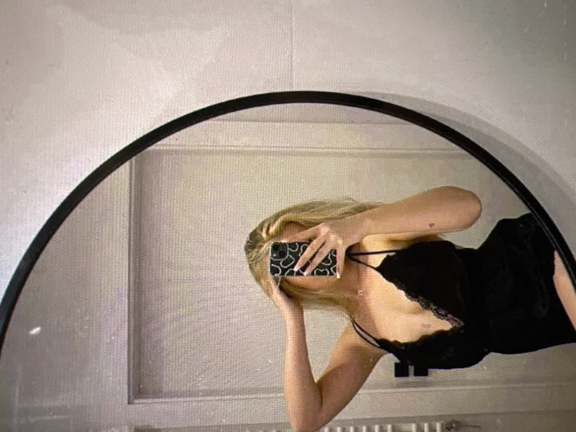 A young woman with blonde hair, wearing a black camisole, is taking a mirror selfie in what appears to be a bathroom. She is holding her phone with a patterned case in her right hand, obscuring her face. Her left arm is raised, with her hand resting on her head. The background shows a white tiled wall, a white radiator, and a door. The image is taken from a slightly low angle. The location in Castelo Branco, Portugal is unknown.