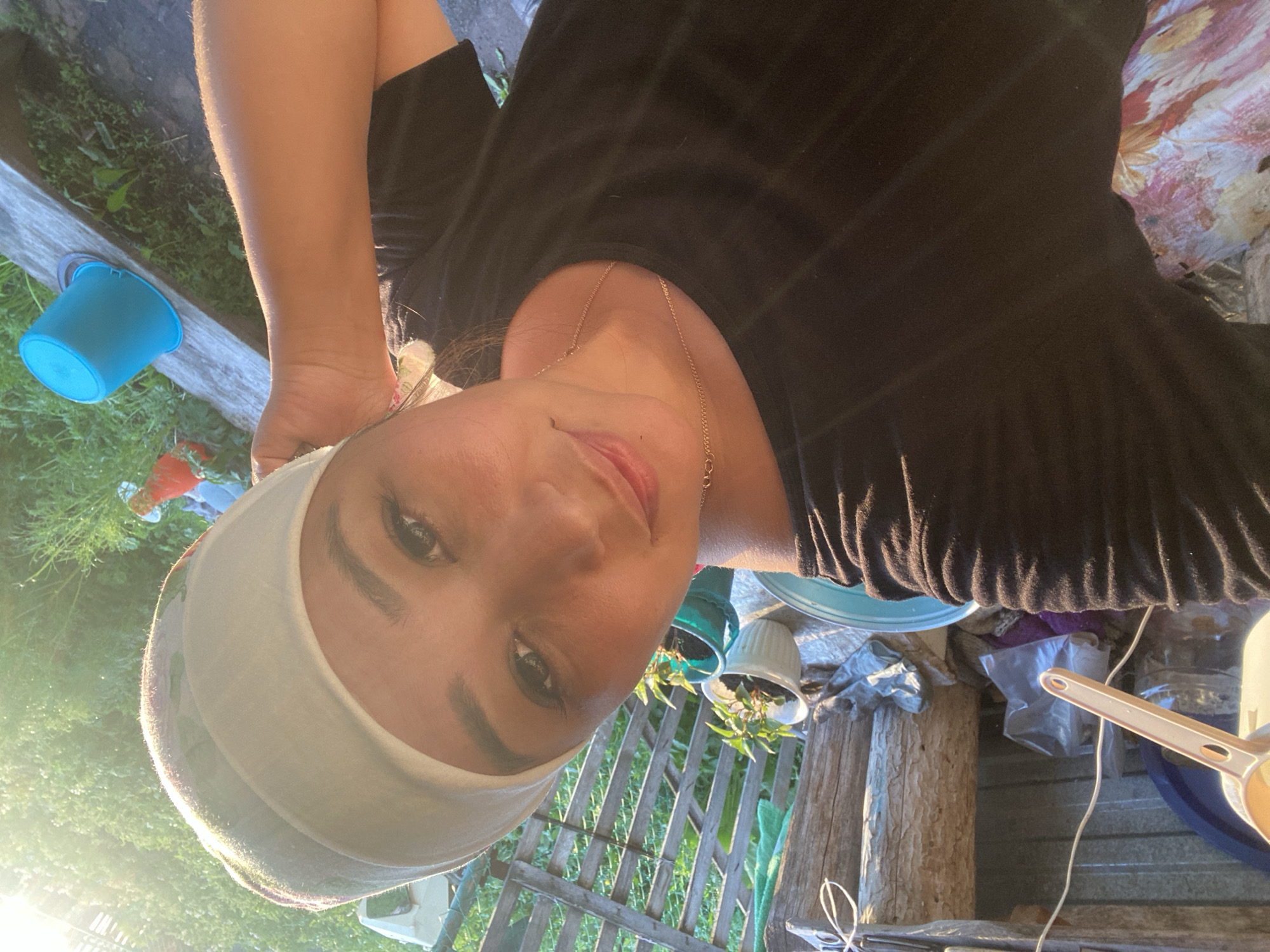 In an outdoor setting that appears to be a rural area or dacha in Maloarslangulovo, Russia, a person with dark hair and wearing a black t-shirt and a white headscarf is captured in a close-up selfie. The person's face is turned towards the camera, with their right arm resting on their head. Sunlight streams in from the right side of the frame, creating a lens flare and highlighting the greenery in the background. Behind the person, various objects are visible, including a wooden fence, several potted plants, a blue bucket, and what appears to be a tarp or some fabric. The overall scene suggests a casual, possibly agricultural or gardening-related activity.