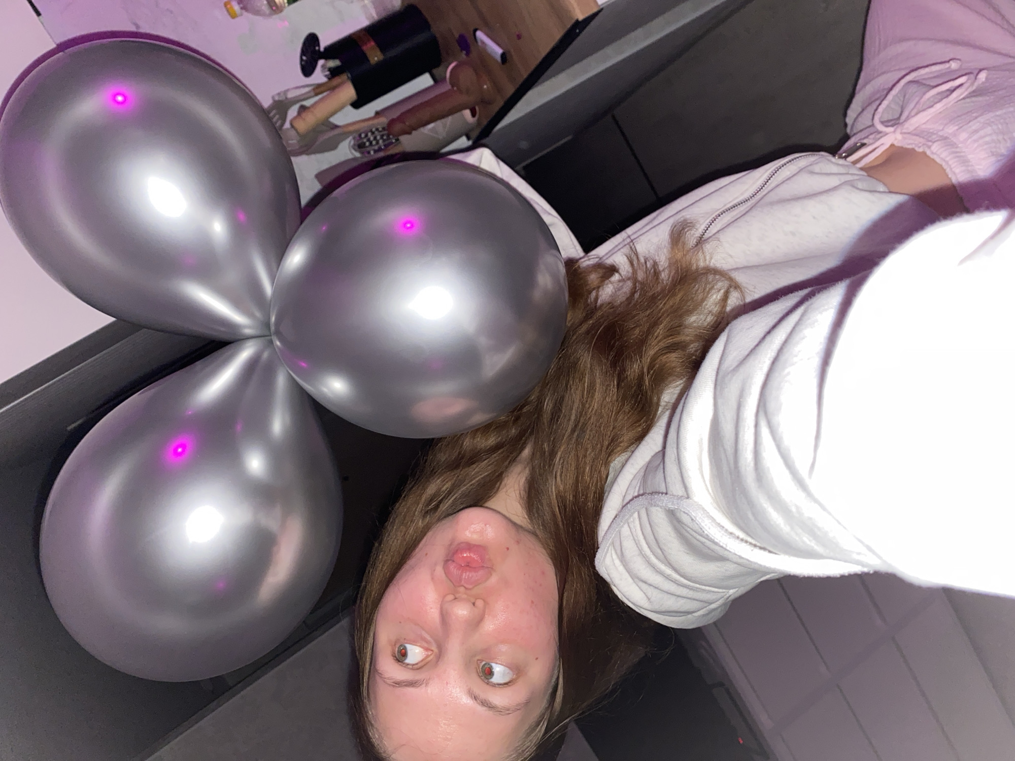 The image is a selfie taken indoors, likely in a domestic setting. A young Caucasian female with long, brown hair is the primary subject, positioned in the foreground. She is wearing a white hooded jacket and light pink pants. Her facial expression is playful, with her lips pursed and eyes looking upwards. She is holding up three silver balloons, two of which are visible in the upper part of the frame, and one in the lower right. The background is dimly lit and appears to be a kitchen or a living area with some objects on a counter, including what looks like a black container with some items inside, a bottle of liquid, and a pen. The lighting suggests an evening or indoor setting, with a slight pinkish hue cast by what might be ambient party lights or reflections. There is no discernible indication of the city of Izhevsk, Russia in the image.