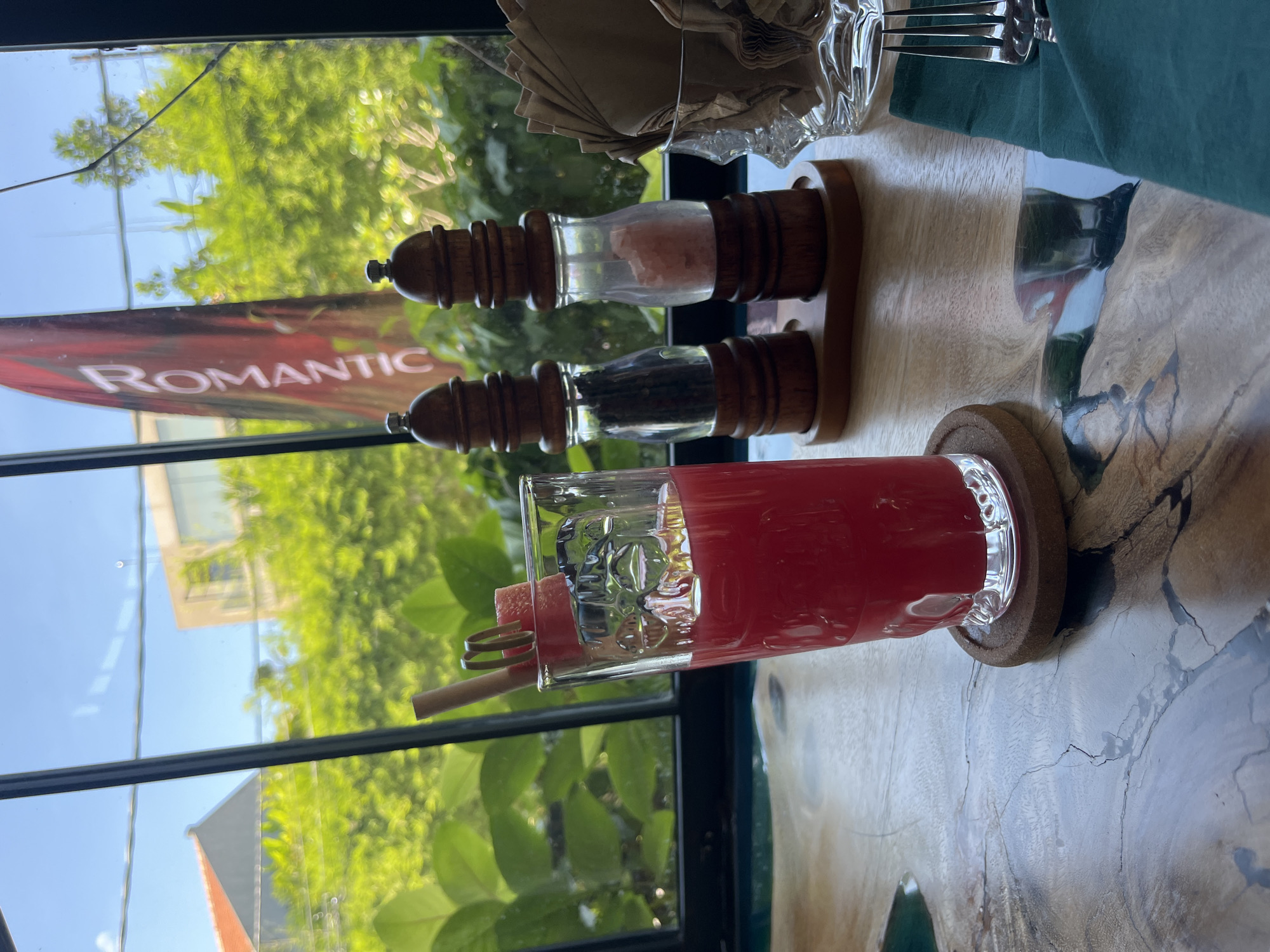 A horizontal, eye-level photograph captures a table setting indoors, with a view of the outdoors visible through a window. The table is set with a pink beverage in a clear glass, decorated with a stick and watermelon. A pair of salt and pepper mills rest on the table, alongside a napkin holder and a fork set. The table's surface has a distinct pattern, resembling wood grain with inlaid blue-green accents.
The window reveals an outdoor scene with greenery and a building visible in the background. A sign with the word "ROMANTIC" is partially visible through the window.
The photograph was taken in North Kuta, Indonesia.