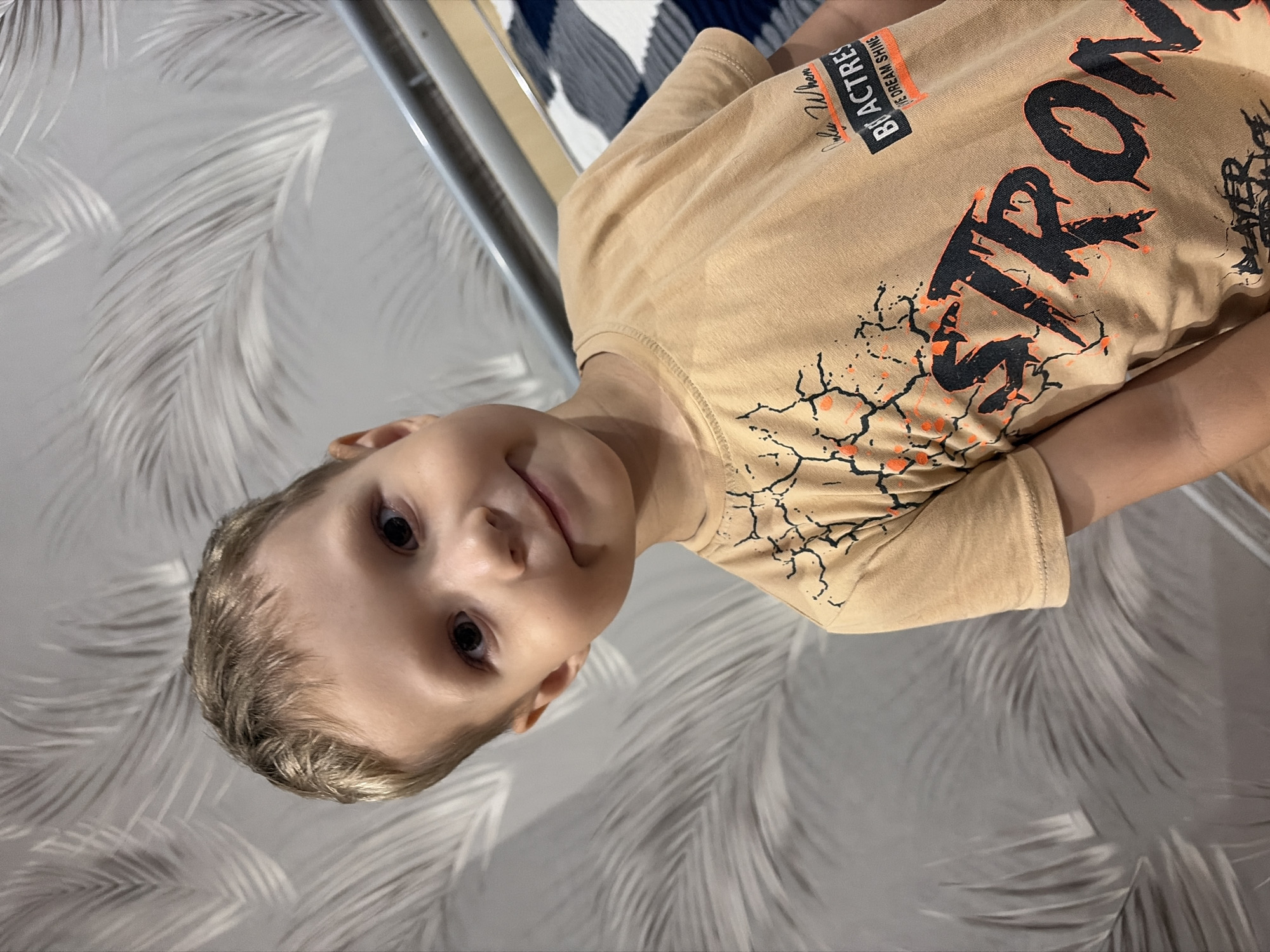 A young boy with blond hair and dark eyes is looking at the camera with a slight smile. He is wearing a light brown t-shirt with a graphic design featuring the word "STRONG" in large, black, distressed letters, along with other text and orange accents. The background consists of a grey wallpaper with a repeating pattern of light grey palm leaves. A metal railing is visible to the right of the boy, and behind it, a blue and white striped blanket or bedding can be seen. The lighting suggests an indoor environment, possibly a home. The image was taken in Chaikovsky, Russia.