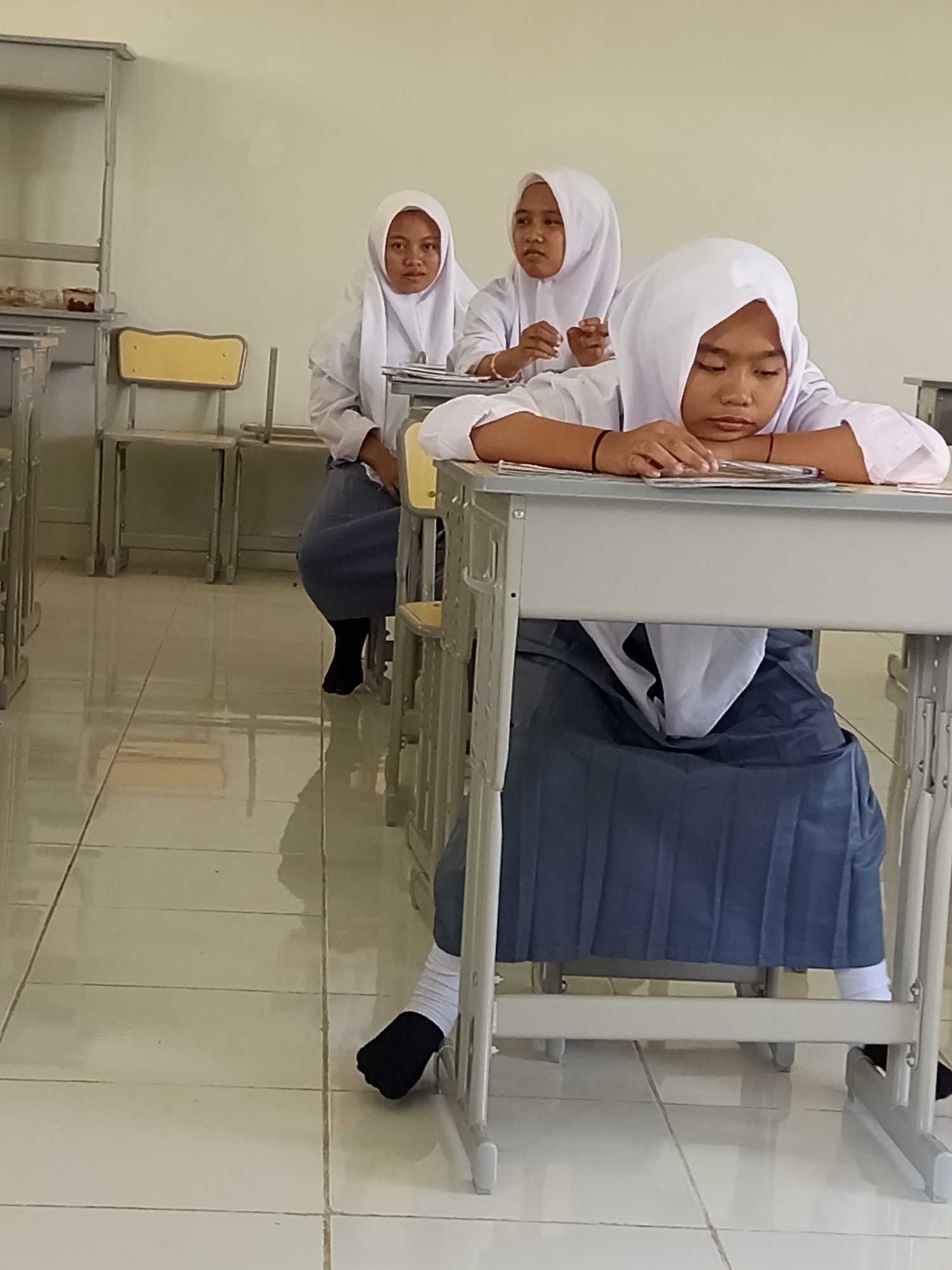 The image depicts three young female students wearing white hijabs and uniforms, seated at desks in what appears to be a classroom. The student in the foreground is resting her head on her arms, which are on a stack of papers, with her eyes closed or looking down. The other two students are positioned behind her, with one looking towards the foreground student and the other looking forward. The classroom has light-colored walls and a tiled floor that reflects light. Desks and chairs are visible, suggesting a school environment. The overall atmosphere suggests a moment of quiet, possibly during a lesson or study period. The image is undated and unlocated.