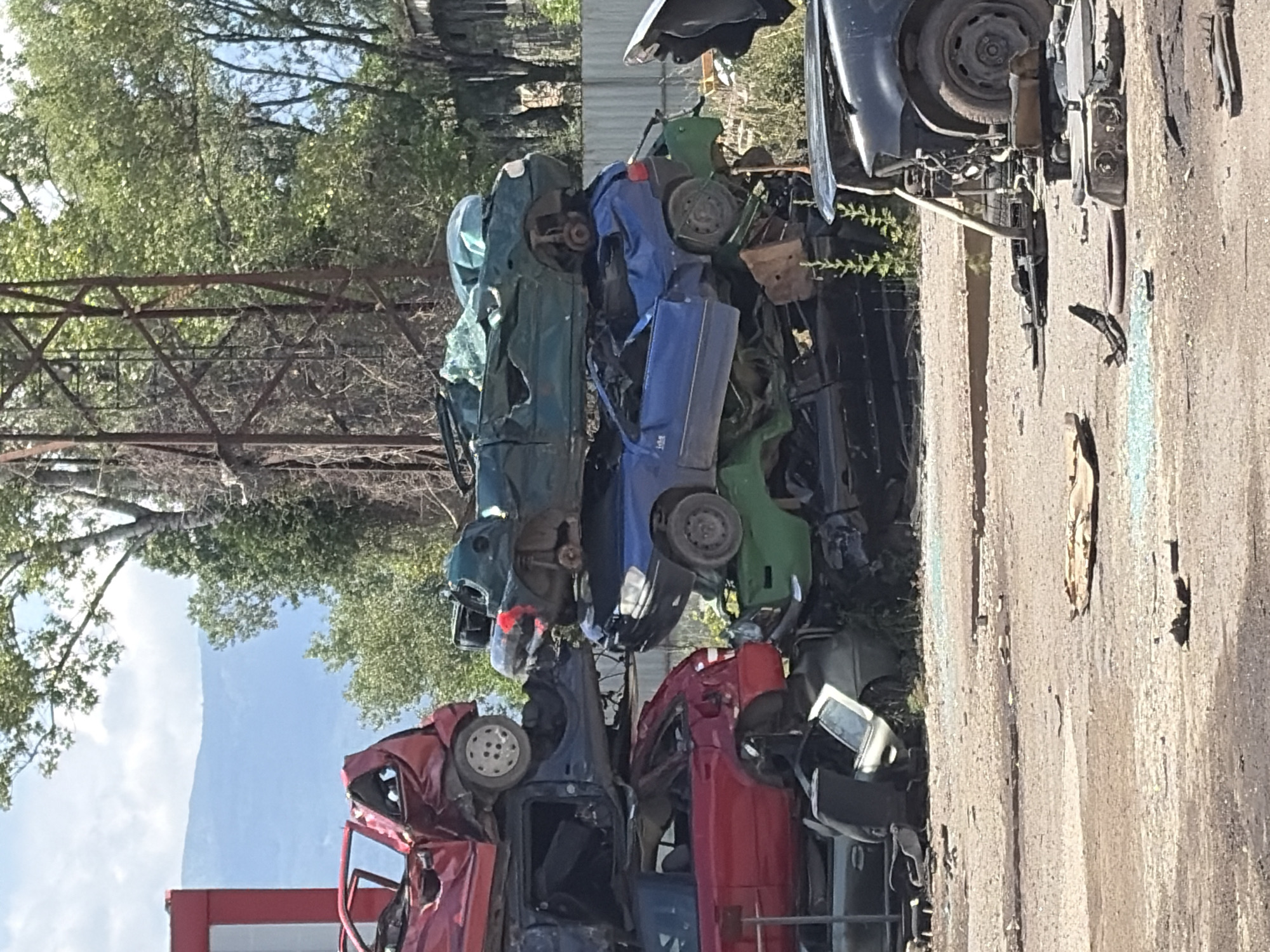 This media file depicts a junkyard filled with a large pile of crushed and stacked cars. The cars are of various colors, including red, green, blue, and dark gray. Some are oriented vertically, while others are tilted or upside down. The scene is outdoors, with trees and foliage visible in the background, suggesting a natural setting juxtaposed with the industrial debris. The sky appears partly cloudy and bright, indicating daytime, likely a sunny day.
The junkyard is an **object** that serves as the **scene** and **setting**. The main **subjects** are the numerous derelict cars, which are **objects** themselves. There are no visible people in the media file. The **activity** is the static state of cars in a junkyard, implying they have been decommissioned and are awaiting processing or disposal.
Notable **details** include the haphazard stacking of the vehicles, highlighting the volume of waste. The texture of the ground appears to be gravel or dirt. The presence of trees and a mountain or large hill in the far background provides a sense of location, possibly rural or on the outskirts of a populated area. The image is oriented vertically, so the scene appears on its side. The overall **emotion** evoked is one of decay and obsolescence, common to scenes of car graveyards. No visible text is present in the image. Given the location context of Busmantsi, Bulgaria, this is likely a car junkyard in that region.