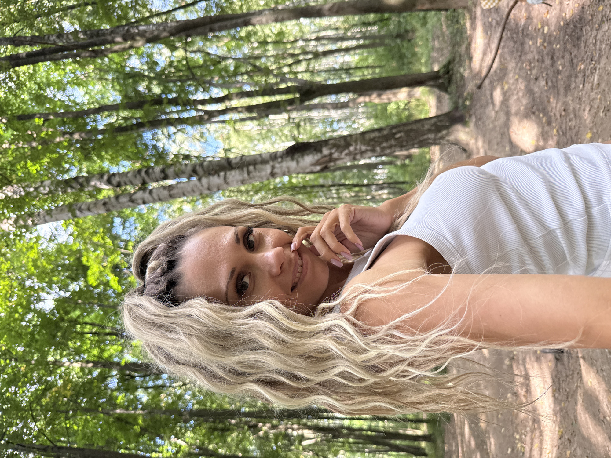 A woman with blonde hair tied in a high ponytail is pictured in a forest. She is wearing a white ribbed tank top and smiling at the camera, with her hand near her mouth. The background shows numerous trees with green foliage and some sunlight filtering through. The ground appears to be covered in dirt and fallen leaves, with a path visible in the distance. The image was taken in the city of Dumino, Russia.
