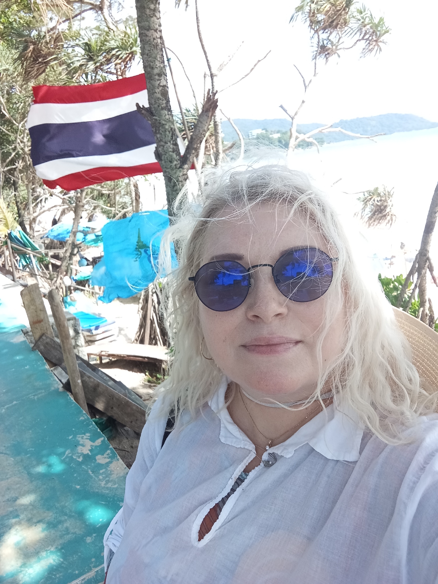 A woman with blonde hair and sunglasses takes a selfie on a beach in Karon, Thailand. She is wearing a white shirt and a straw hat. In the background, the Thai flag is visible, along with palm trees and the ocean. The scene appears to be during the day, with bright sunlight.