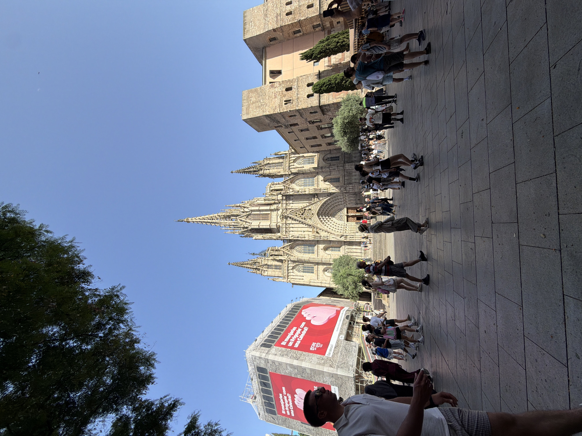 This media file depicts a lively scene in Barcelona, Spain, likely during the daytime given the bright sunlight casting distinct shadows. The primary subjects are a large gathering of people walking on a paved plaza in front of a magnificent Gothic cathedral. The cathedral, with its intricate spires and detailed architecture, dominates the background, suggesting a significant historical or religious landmark.
Several individuals are captured in mid-stride, suggesting movement and a bustling atmosphere. The people are dressed in casual summer clothing, indicating warm weather. The composition offers glimpses of various architectural styles, including a more modern building to the left adorned with large, red banners. One banner prominently features text, partially legible as "El menjaries un Frigopie com una Catedral?", which appears to be an advertisement. Another banner below it repeats a similar slogan.
The scene appears to be a popular tourist destination, with many people likely visiting the cathedral or exploring the surrounding area. The clear blue sky above further reinforces the impression of a pleasant day. The overall mood seems to be one of leisurely exploration and engagement with the urban environment. The presence of trees and the tiled pavement contribute to the urban setting, while the grandeur of the cathedral provides a focal point.