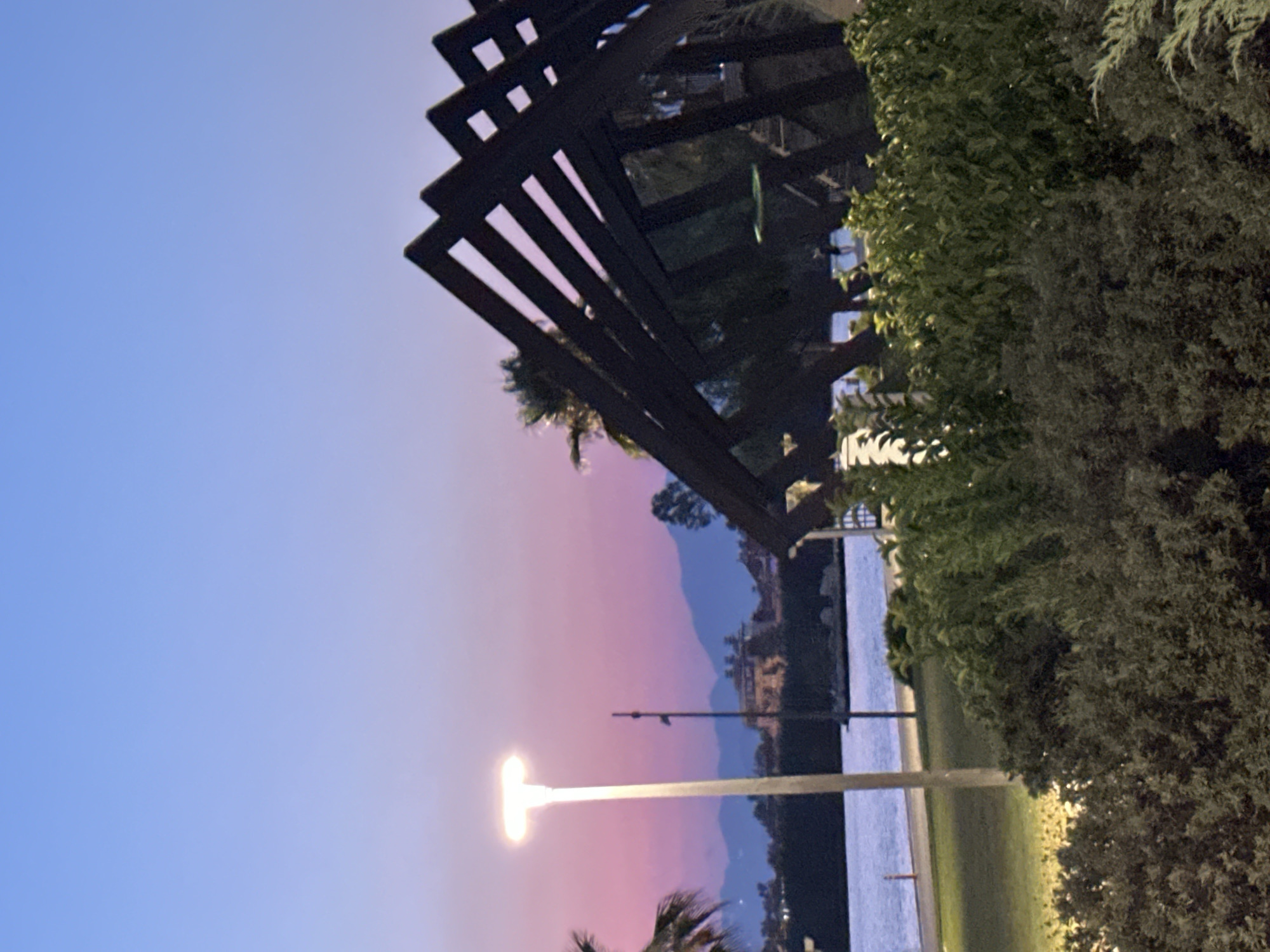 The image depicts an outdoor scene at dusk in Fethiye, Turkey. In the foreground, lush green bushes and some evergreen shrubs are visible, with a park lamp on the left side of the frame. In the middle ground, a body of water, likely the Mediterranean Sea, is present, with a pathway and some distant buildings and trees on the opposite shore. The background features silhouetted mountains under a twilight sky, transitioning from a soft pink hue near the horizon to a clear blue overhead. A modern, angular wooden structure is prominent on the right side of the image. There are no discernible people or actions taking place in the frame.