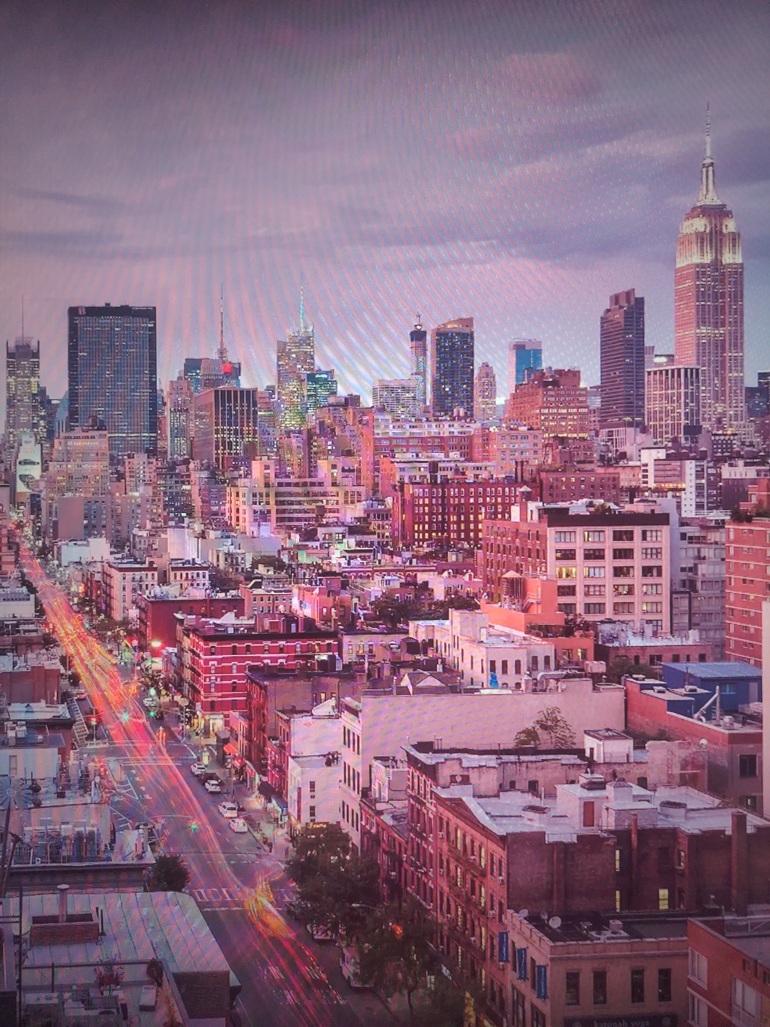 The image is an aerial cityscape at dusk with a prominent display of tall buildings, suggesting an urban center, likely New York City due to the presence of the Empire State Building on the right. The city is characterized by high-rise structures, with dense clusters of buildings in the foreground and extending into the distance. Street-level movement is visible, evidenced by light streaks indicating vehicular traffic on streets, the location in the city of Unknown, Unknown. The sky is overcast with a pinkish hue from the setting sun, adding a warm tint to the scene. Buildings vary in height and architectural style, some with traditional brick facades and others with modern glass structures.