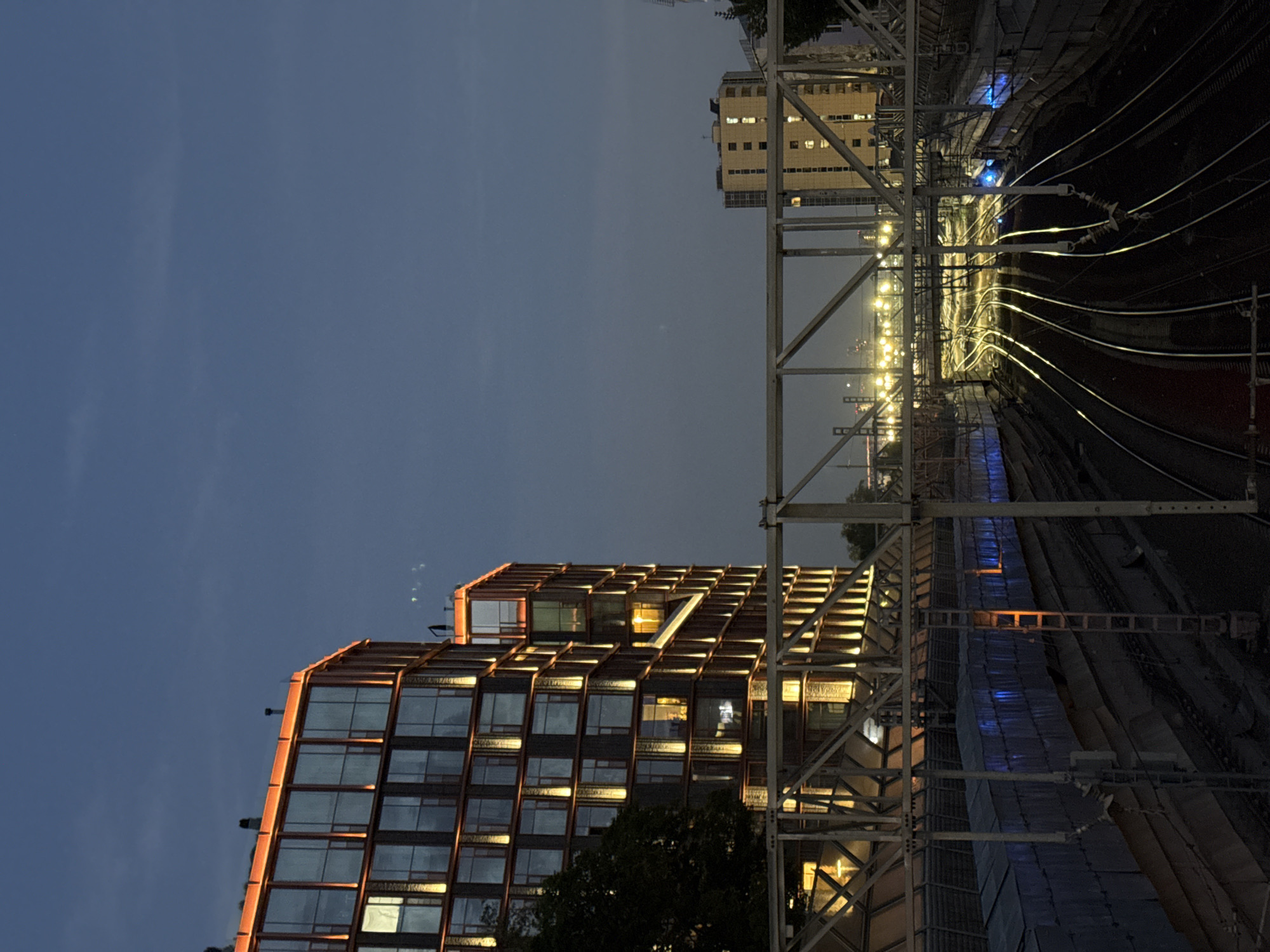The image depicts a twilight view of a city street in Moscow, Russia. In the foreground, multiple train tracks curve through a somewhat industrial area. A pedestrian bridge with a metal railing spans across the tracks. On the left, a modern building with a glass facade is illuminated by warm orange lights along its edges and vertical strips. The building has many windows, some of which are lit from within. To the right of this building, a taller, lighter-colored building with many windows is visible in the distance. The sky is a deep blue, characteristic of dusk.