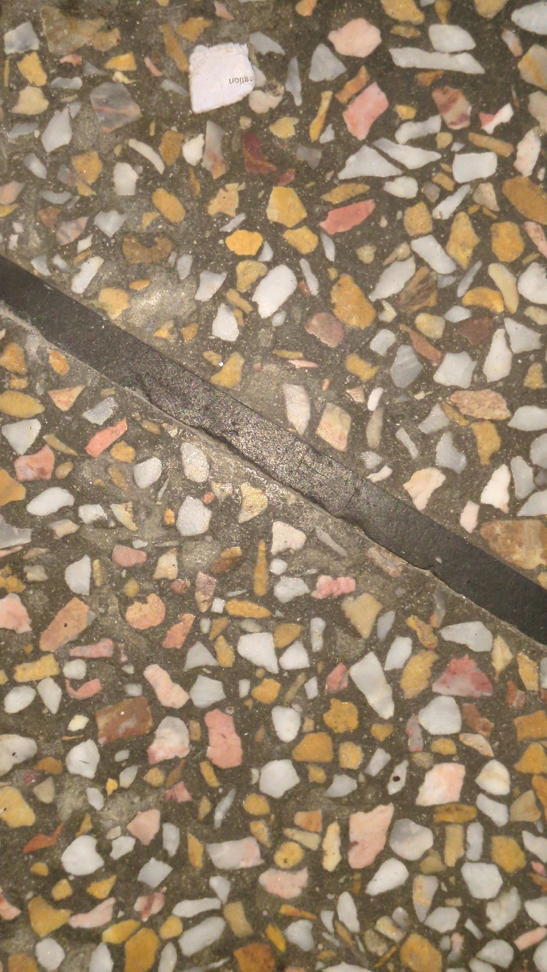 The image shows a close-up, overhead view of a speckled terrazzo floor. The floor is composed of a dark gray base material with numerous irregularly shaped chips of white, pink, and yellow aggregate embedded within it. A thick, dark gray, textured seam runs diagonally across the image, dividing the floor into two sections. A small, torn piece of white paper with black text that appears to be "ration" written vertically is visible on the upper left quadrant of the floor. The lighting is even, and the surface appears slightly wet or polished, reflecting some light. The context of Grass Mandi, Pakistan is not visually inferable from the image itself.