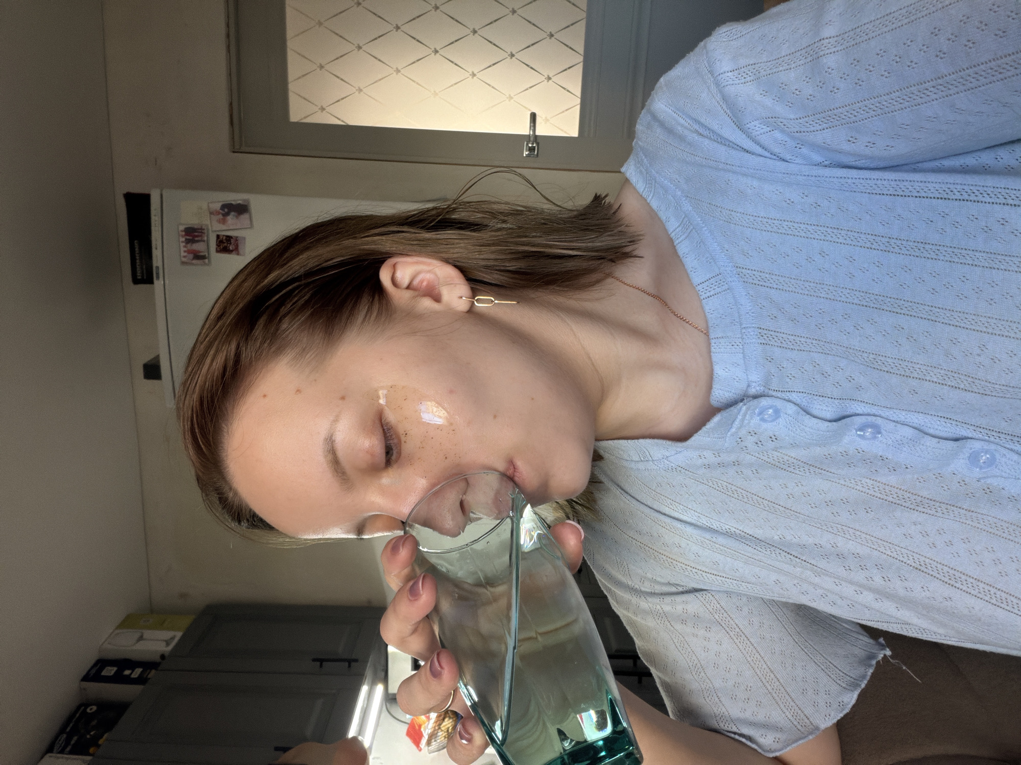 A woman is drinking from a glass. She is wearing a light blue, buttoned shirt, and gold earrings. The location appears to be indoors, possibly a kitchen. A refrigerator with magnets is partially visible in the background. The location is in the city of Reshetnikovo, Russia is not verifiable from the provided image.
