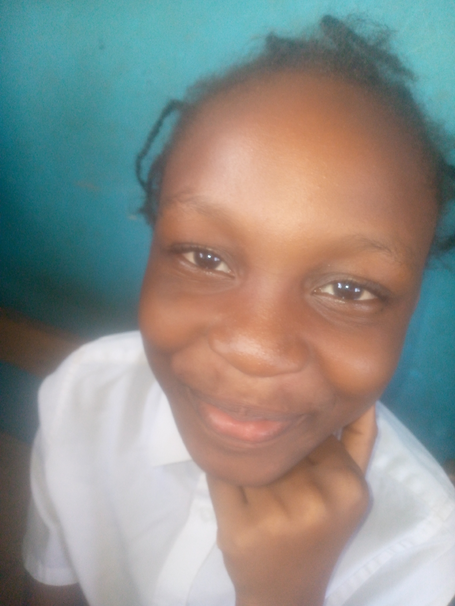 A young Black girl with dark hair pulled back from her forehead smiles gently at the camera, resting her chin in her hand. She is wearing a white collared shirt. The background is a blurred blue. The location of the photo is Ikorodu, Nigeria.