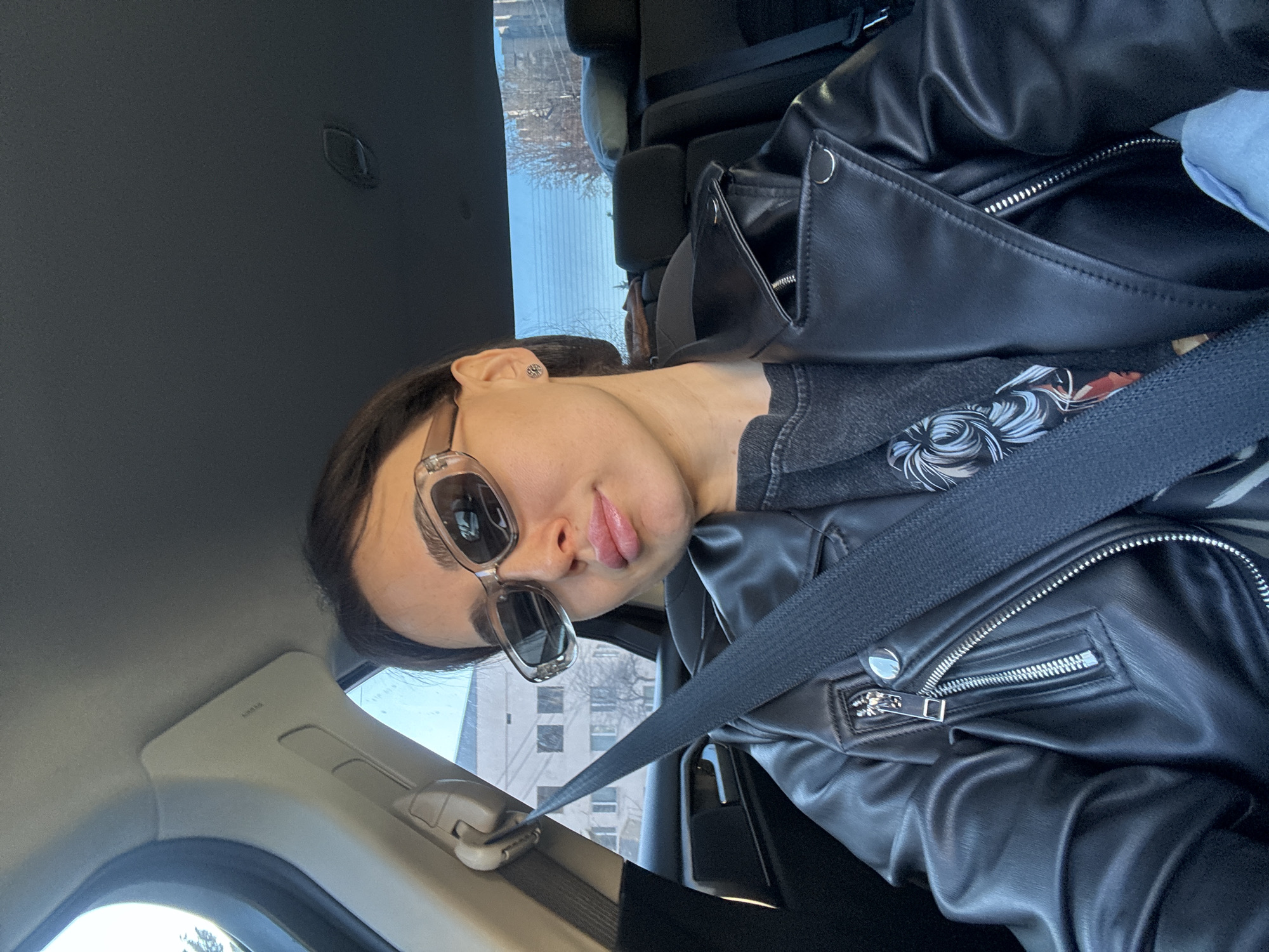 A woman is seated in the driver's seat of a car, wearing a black leather jacket, sunglasses, and a seatbelt. The car is in Tbilisi, Georgia, as evidenced by the architecture visible through the window. The focus is on the woman's face and upper body.