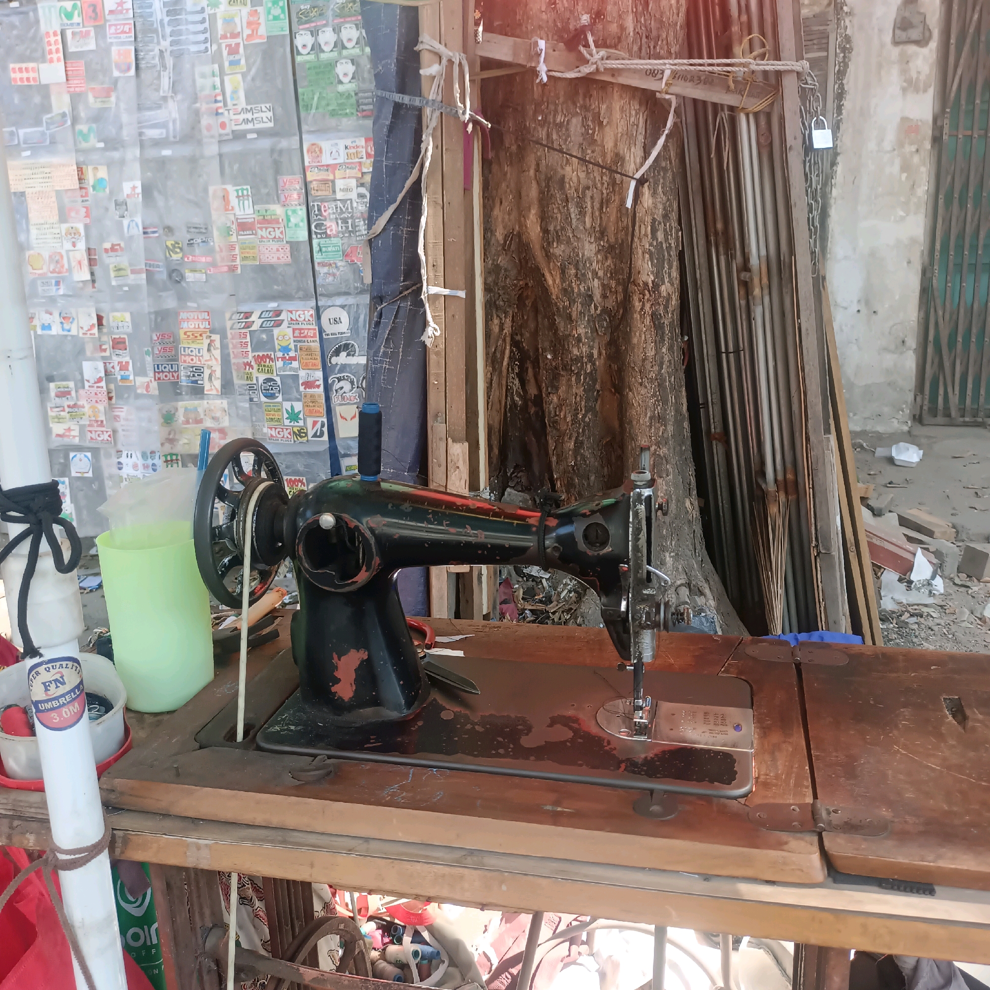 A vintage black sewing machine with red accents sits on a worn wooden table. The machine has a large metal wheel on the left and a spool of black thread on top. A green plastic cup and a white tube with black rope are next to it. Behind the sewing machine, a large tree trunk is visible, with various wooden planks and metal poles leaning against it. A wall covered in stickers and posters is to the left of the tree. The scene appears to be an outdoor workspace in Surabaya, Indonesia, potentially a small shop or market stall. The overall impression is one of a functional, albeit cluttered, environment.