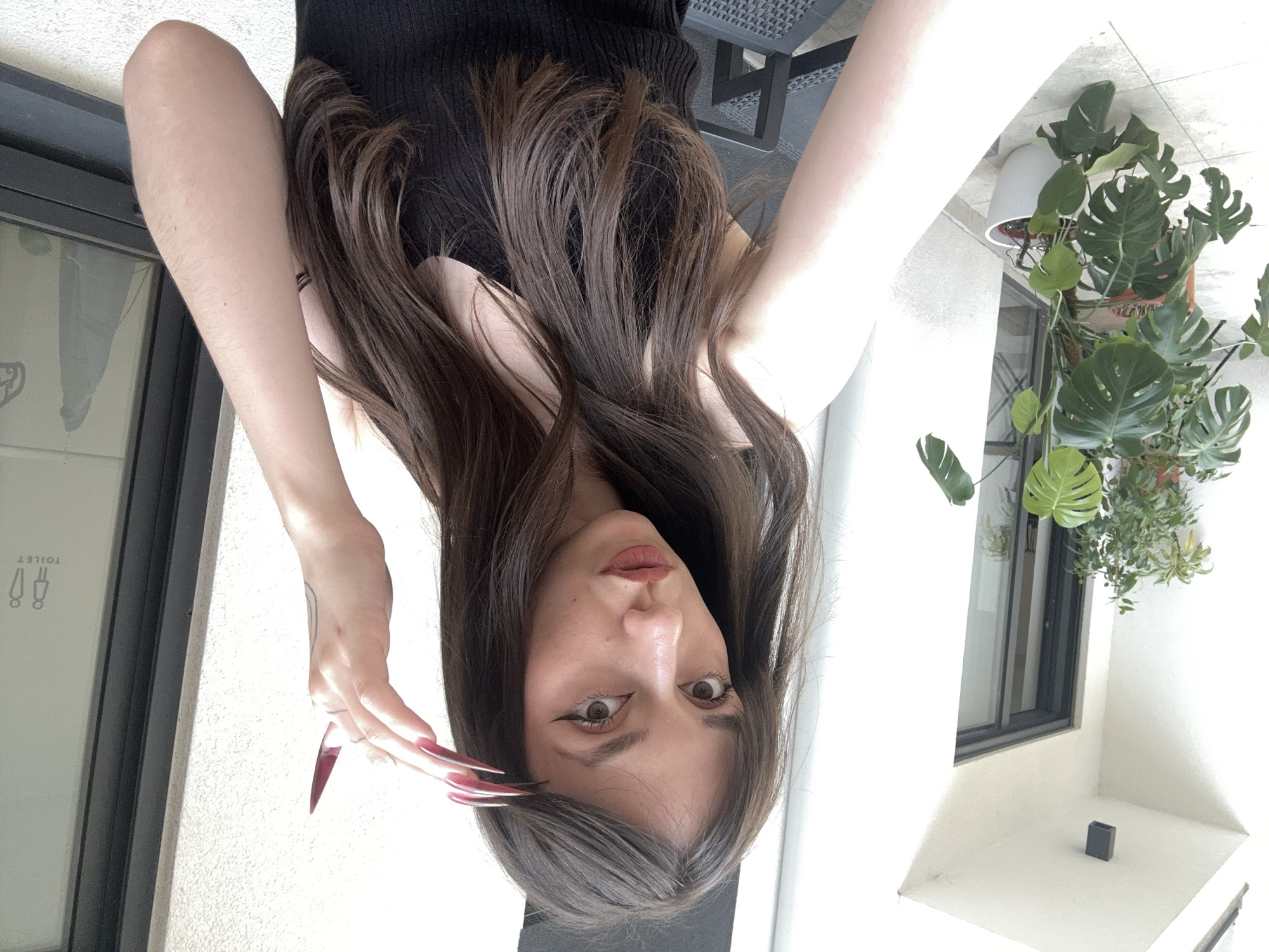 A young woman with long, dark hair, and long, pointed, red nails is posing for a photo. She is wearing a black top. Behind her is a white wall with a window. In the window are green plants. To the left of the woman is a dark wall with a toilet sign. The location is likely in Vracar Urban Municipality, Serbia, based on visual clues and context. The woman is the main subject of the photo.