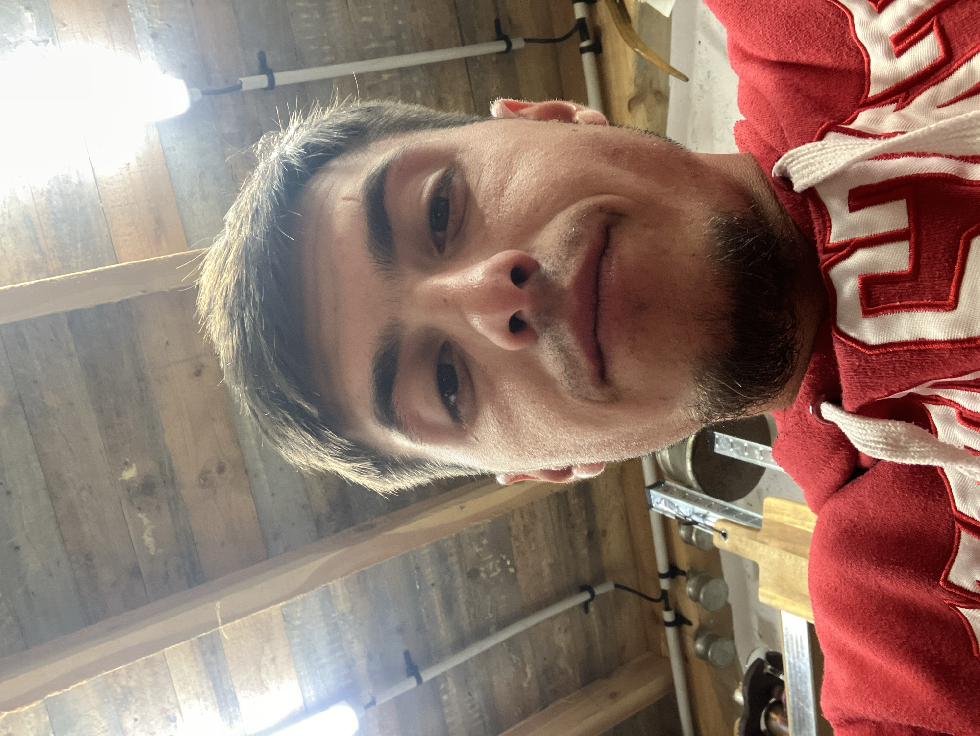 A man is taking a selfie in what appears to be a workshop or garage. The background consists of wood-paneled walls and a visible lighting fixture. To the right of the frame, parts of a red hooded sweatshirt are visible. There is no evidence of the location being in Komuniga, Bulgaria.
