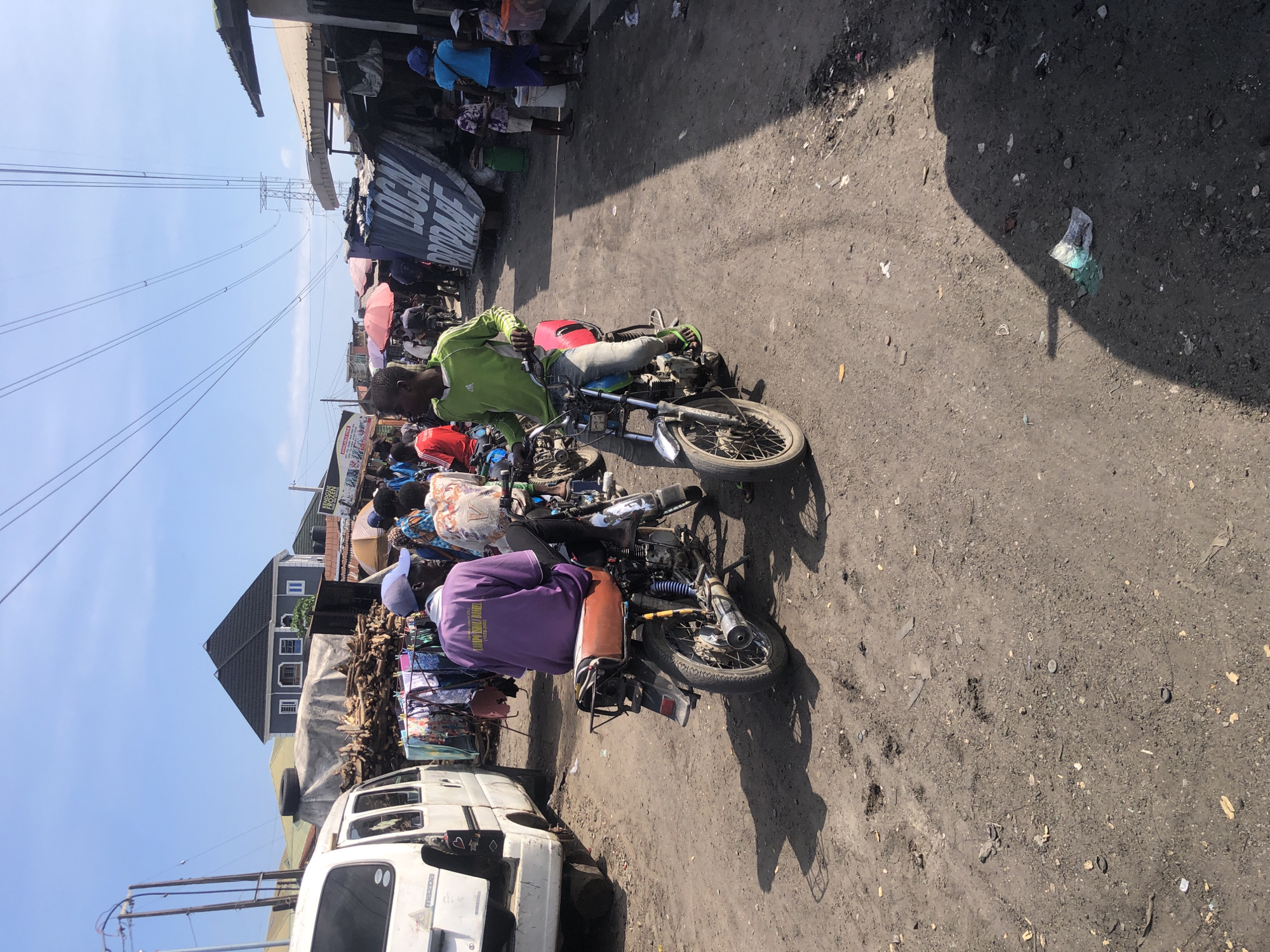 The image depicts a bustling outdoor market scene in Ado, Nigeria, under a clear blue sky. The foreground is dominated by a dusty, unpaved road. Two young men are prominently featured, each on a motorcycle. The man on the left is wearing a purple t-shirt and a blue cap, seated on a motorcycle with a brown seat and yellow shock absorbers. The man on the right is wearing a green long-sleeved shirt and jeans, riding a motorcycle with a red seat and chrome handlebars.
Behind them, the market stretches out. To the left, a white van, possibly a Mazda, is parked. Piles of chopped wood are visible, covered by a large tarp. Clothes hang on a line between stalls. Various market stalls with awnings and umbrellas are set up, suggesting a lively commercial area. In the background, a modern-looking building with a dark pitched roof stands out. Overhead, power lines crisscross the sky, supported by transmission towers. The ground is littered with debris, including leaves, plastic wrappers, and scattered objects, indicating a well-used and active public space. The overall impression is one of vibrant daily life in a Nigerian urban market environment.
