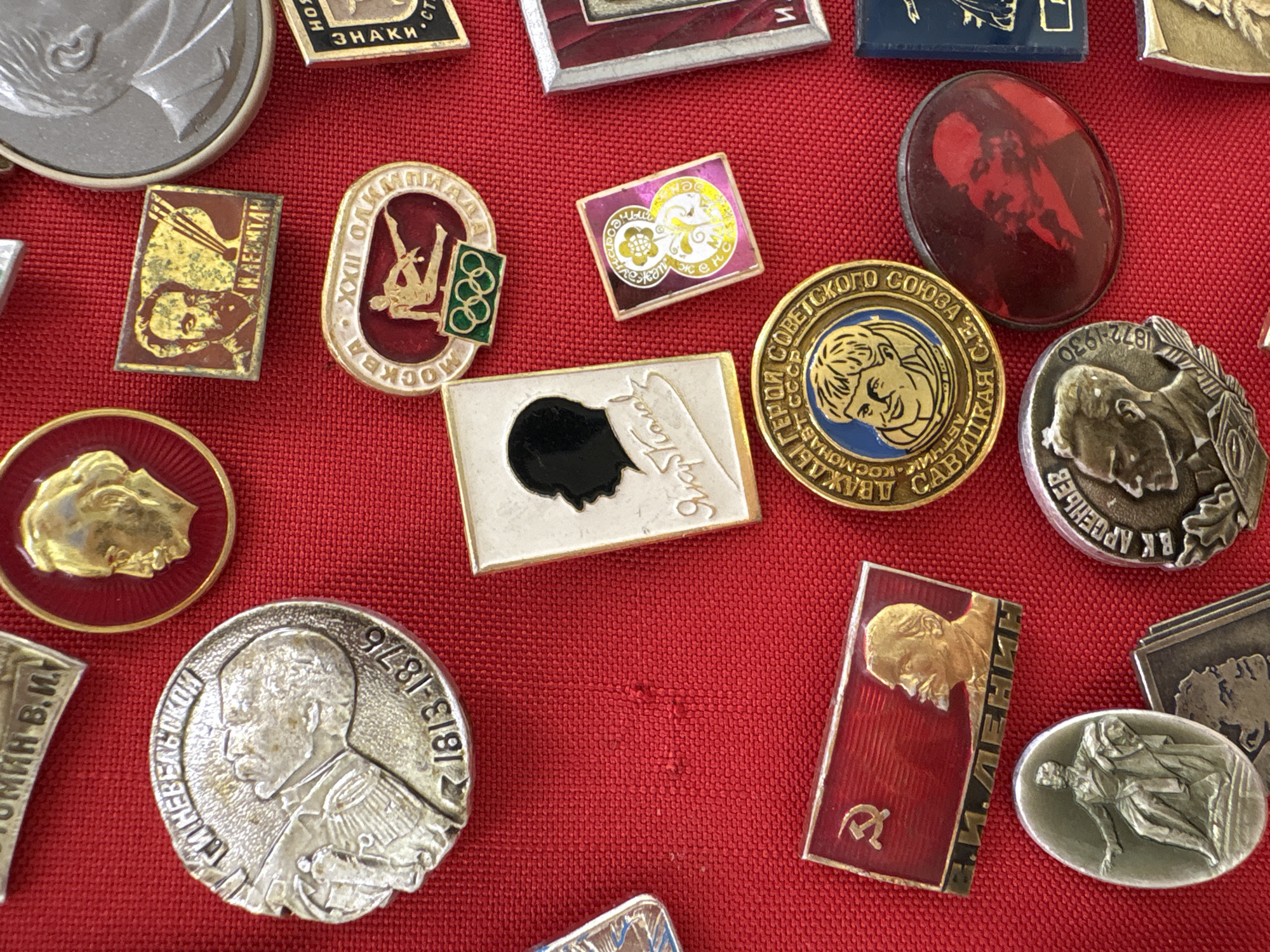 The image displays a collection of Soviet-era badges and medals arranged on a textured red fabric surface. The objects vary in shape, size, and color, featuring portraits of historical figures, symbols, and text in Russian. The background is a vibrant red with a visible fabric texture. The collection appears to be laid out flat for display or organization, with no people or actions depicted. The overall impression is one of historical artifacts. No specific location in Moscow, Russia is evident from the image itself.