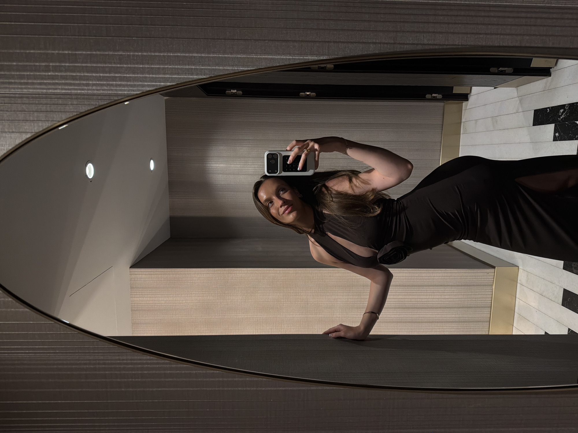 The image depicts a female in Budva, Montenegro, taking a mirror selfie indoors. The woman, with long dark brown hair, stands to the left of the frame, facing the arched, gold-framed mirror that dominates the left half of the composition. She is wearing a dark, possibly black, form-fitting halter-neck gown featuring a keyhole cutout on the chest and a decorative dark rosette embellishment at the waist on her left hip. A high slit is visible on the left side of the dress. Her left hand, adorned with a thin gold-colored bracelet, rests against a vertically striped, light beige wall panel. Her right hand holds a light-colored smartphone (white or light grey case with dark buttons/features) at chest level, pointed towards the mirror. She is looking at the phone with a slight smile. A gold-colored ring is visible on her right ring finger.
The environment is a luxurious interior space, likely a hotel hallway. The walls feature vertical striped wallpaper in varying shades: a darker grey/brown to the immediate right of the mirror and a lighter beige/tan on the panels she is leaning against and to their right. Gold-colored metallic baseboards run along the bottom of the wall sections. The floor is tiled with a contrasting black and white pattern, consisting of both plain white and black marble-patterned tiles arranged geometrically.
The mirror's reflection shows a white ceiling with multiple recessed circular lights and a thin black linear vent. On the right side of the mirror's reflection, dark doorframes with gold-colored hardware (hinges/handles) are visible, reflecting a continuation of the vertically striped wallpaper pattern. The lighting is dim, casting soft shadows and highlighting textures.
