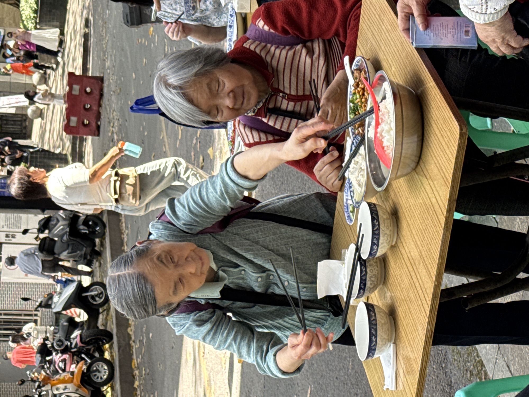 Two elderly women sit at a light-wood rectangular table outdoors, using chopsticks to eat from bowls. The woman on the left wears a light blue sweater and has short grey hair. The woman on the right wears a red and white striped sweater with a purple shoulder bag strap. Dishes of food, including rice and vegetables, are placed on the table between them. The table is set with three bowls, two of which are stacked, and napkins. The area behind the women shows a street scene, including motorbikes and pedestrians. A red traffic barrier is visible. The location is indicated to be in the Yuzhong District of Chongqing, China.