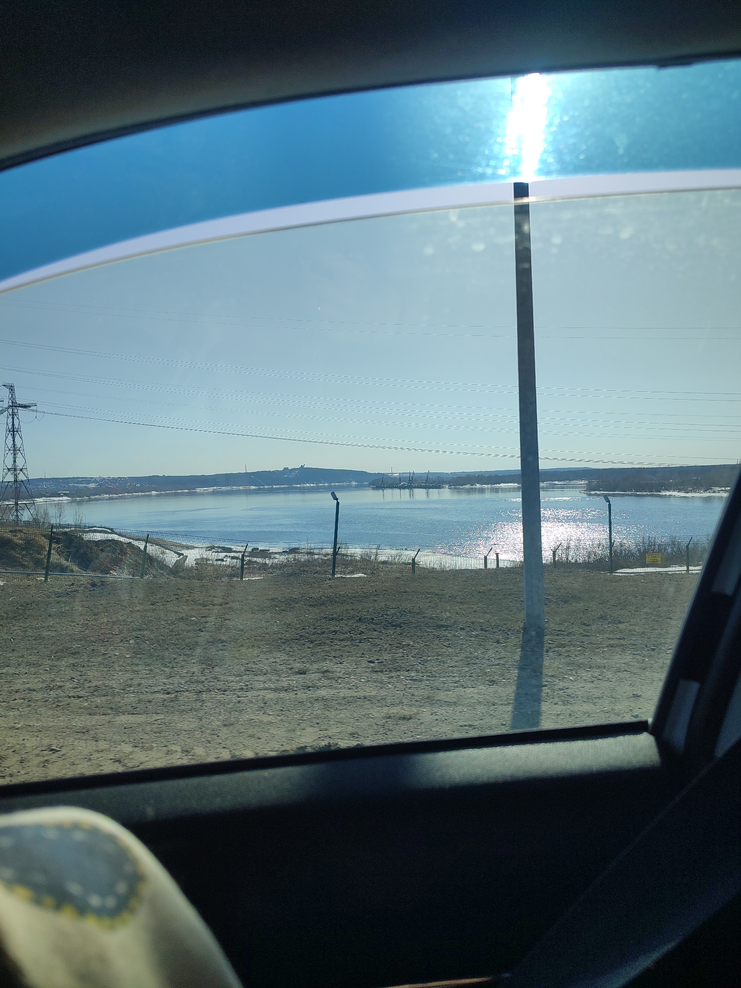 The image, taken from inside a vehicle, shows a view of the Kama River near Chaikovsky, Russia. Through the car window, we see a landscape with a river reflecting sunlight, a sandy or gravelly foreground, and a power line support tower on the left. In the distance across the river, there is a built-up area or structure atop a hill. The immediate foreground includes the car's interior, particularly the window frame and a portion of a seat. A fence runs along the near bank of the river. The sky is a clear blue. The image suggests a daytime, sunny condition.