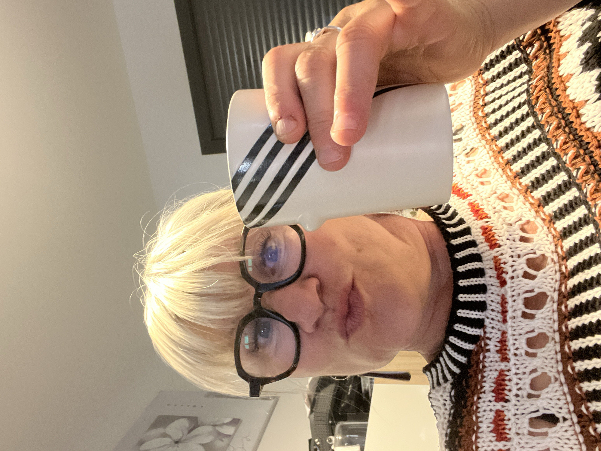 A woman with blond hair and glasses is holding a white mug with black stripes. She is wearing a patterned sweater. The background includes a white wall, a window with closed blinds, and a picture frame. The image was taken indoors, and the specific location in Chambœuf, France cannot be determined from the image.