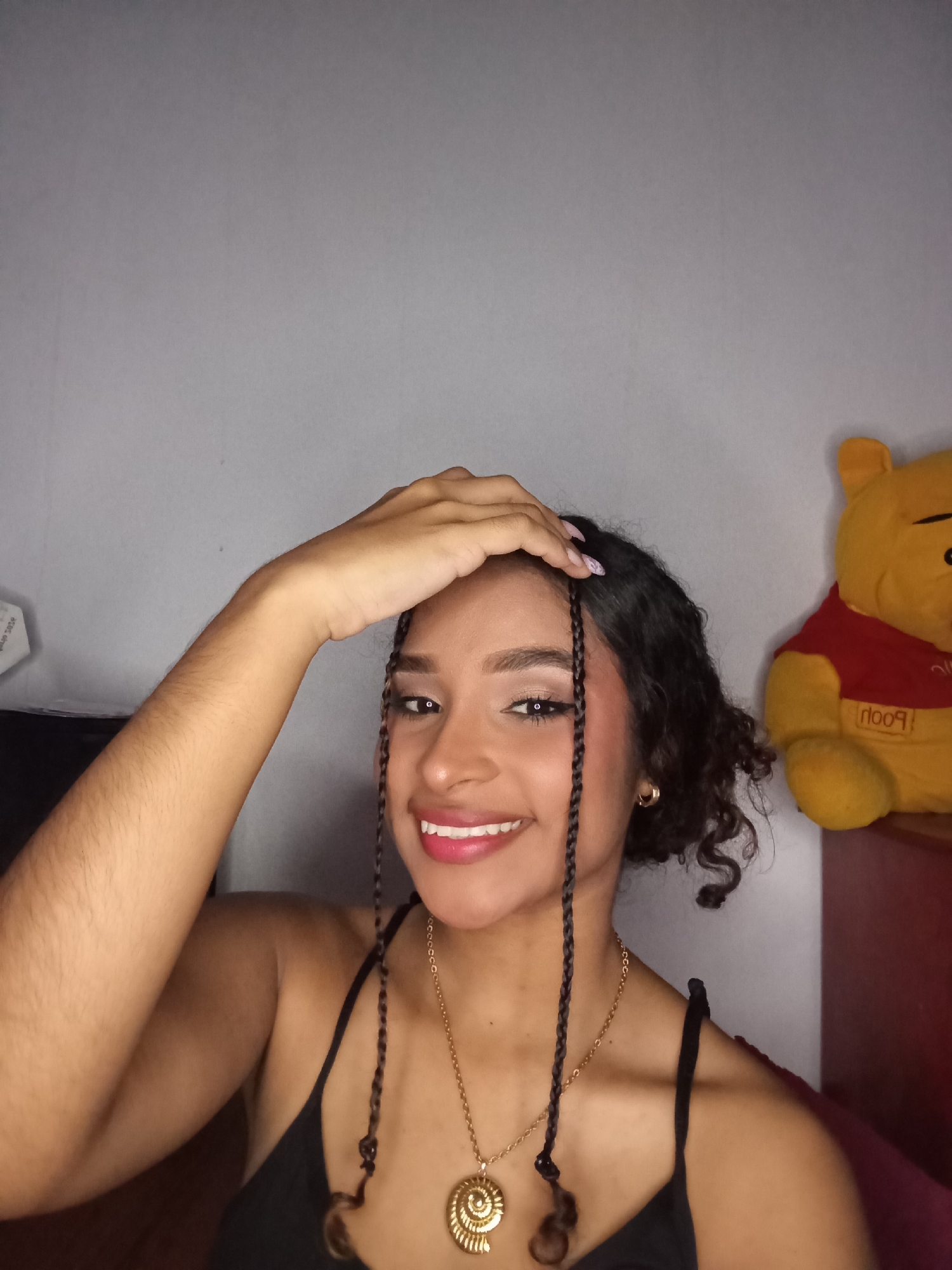 An indoor photograph depicts a young adult female, likely in Juan Griego, Venezuela. The subject has light brown skin, dark wavy hair styled with several small braids framing her face. She is smiling broadly, revealing white teeth, and gazing directly at the viewer. Her right hand is raised, with fingers resting on the top of her head. She wears a black spaghetti-strap top, a gold chain necklace with a large spiral-shaped pendant, and small gold hoop earrings. Her fingernails are painted with a light-colored polish. The background consists primarily of a plain light grey wall. To the right, a yellow Winnie the Pooh plush toy, wearing a red shirt with "Pooh" printed on it, is partially visible, positioned on a dark red surface. A white rectangular object is partially visible on the far left. The scene is illuminated by what appears to be frontal, even lighting.