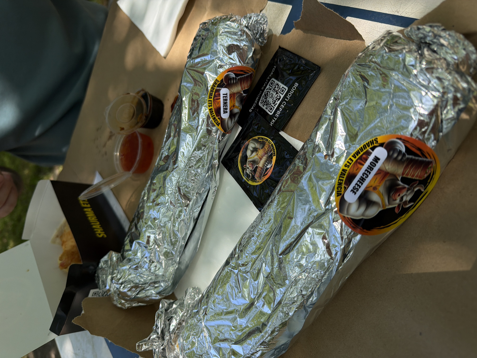 The image shows two foil-wrapped food items in a cardboard box, labeled "SHAURMA ETS VALENCIA," suggesting a location in Valencia, Spain. The food items appear to be shawarmas. Each shawarma has a sticker indicating the filling: one is labeled "TERNERA" (beef), and the other "MORECHEESE" (more cheese). Alongside the shawarmas are wet wipe packets ("TOALLITAS HUMEDAS" and "ВОЛОГІ СЕРВЕТКИ"). There are also small plastic containers with dipping sauces, and a separate white box with a black logo, also labeled "SHAURMA ETS VALENCIA". A person wearing a green shirt is visible on the left of the frame. The items are placed in what appears to be an outdoor setting.
