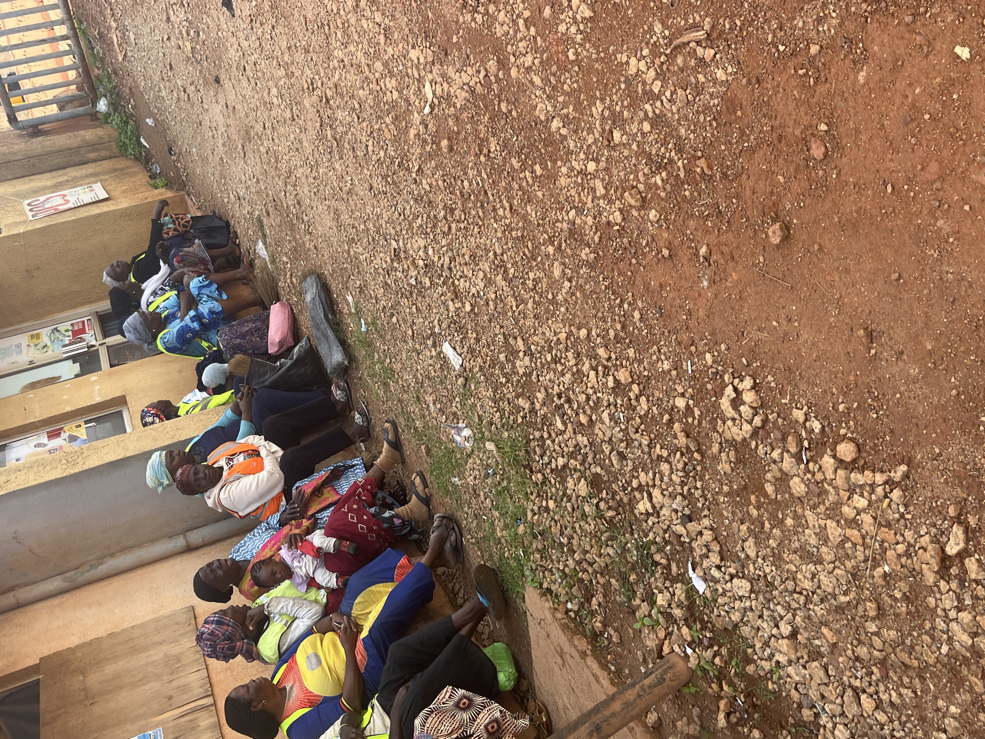 In Gura Top, Nigeria, a group of women are seated on a low concrete barrier outside a building, with their legs extended forward. They are waiting, some are looking to the side and some are looking straight ahead. The ground is covered with gravel and dirt. To the right of the frame, a metal fence is visible. There are some small plants growing around the area. The building has a light brown facade.