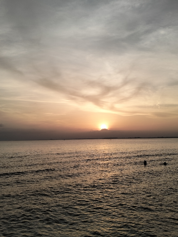 A serene sunset over the water. The sun is partially obscured by clouds on the horizon, casting a golden glow across the water and sky. Gentle waves ripple across the surface of the sea, reflecting the warm light. The sky is filled with soft, muted colors ranging from gray to yellow. Two dark objects are visible in the water towards the right of the frame.