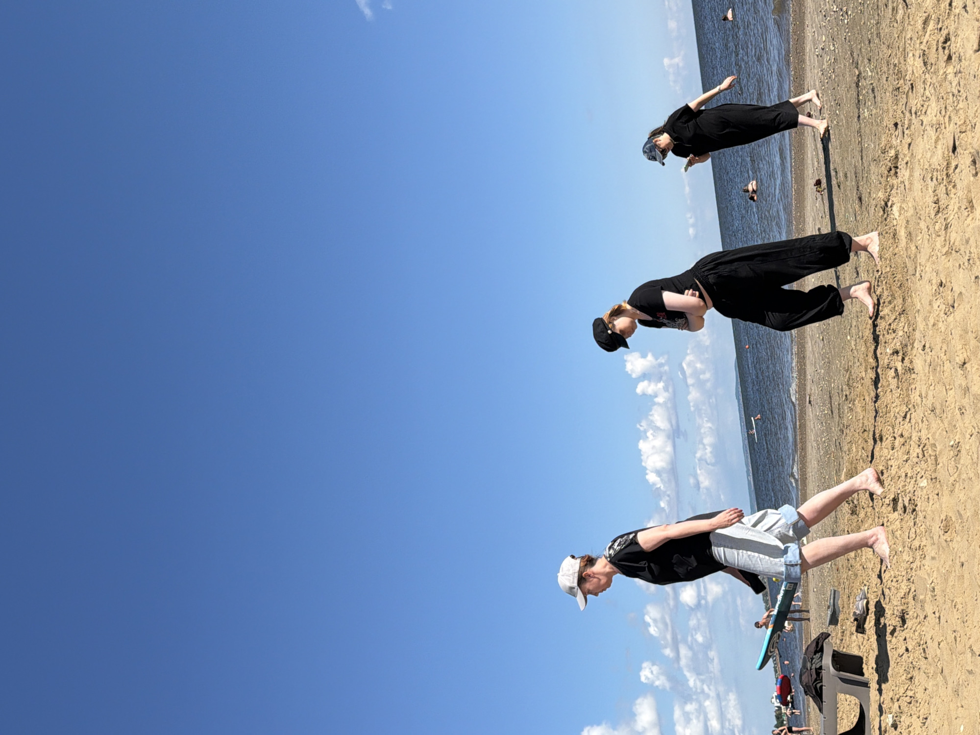 An eye-level, full shot captures three individuals on a sandy beach under a bright blue sky dotted with white clouds. The woman on the left wears a black t-shirt, light blue denim shorts, and a white baseball cap. She is walking to the left, carrying a teal paddleboard under her right arm. A dark object, possibly a bag, rests on the sand to her left. In the middle, a person in a black t-shirt and black baggy pants with a black cap walks to the right, hands clasped in front. To the right, another individual in a black dress and a blue baseball cap walks to the right, looking down at a cell phone in their hand. The ocean with visible waves stretches across the middle ground, with several people swimming or wading in the water. In the background, a distant landmass or coastline is visible. The sand is textured with footprints and small debris.