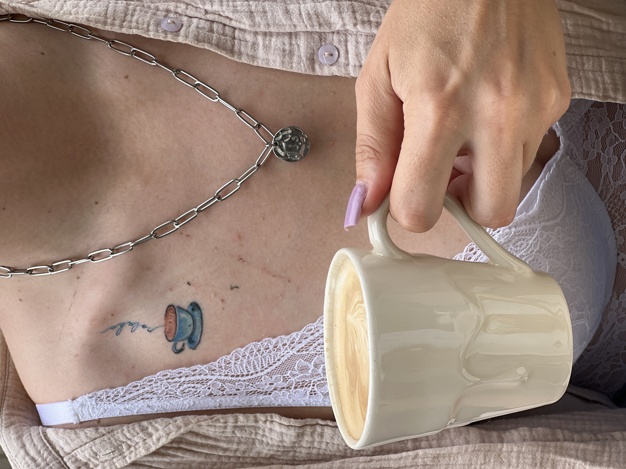 A close-up shot depicts a woman's torso and hand, holding a ceramic mug filled with a light-colored beverage, presumably coffee or latte art. The woman is wearing a light pink button-up shirt with a white lace bralette visible beneath. She has a silver chain necklace and a tattoo of a teacup with accompanying script on her chest. Her fingernails are painted a light purple color. The setting is indoors; I cannot determine if this image was taken in Podgorica, Montenegro.