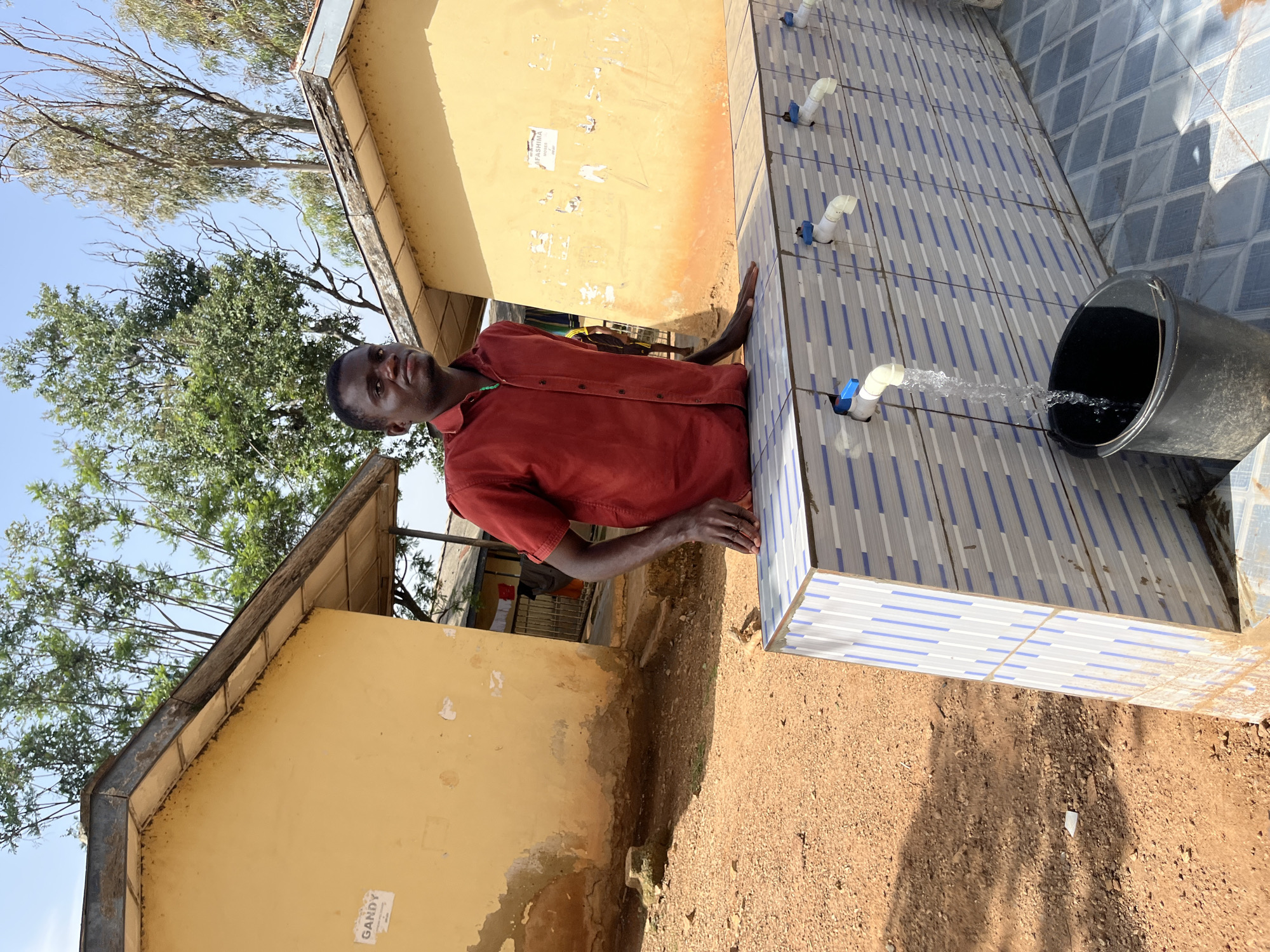 In a sunny outdoor environment, a young Black man wearing a red short-sleeved collared shirt stands with his left arm resting on a tiled surface with multiple faucets, one of which is running water into a black bucket. The man is looking towards the camera. Behind him, two single-story yellow buildings with corrugated metal roofs are visible, along with trees and a clear blue sky. The ground is covered in reddish-brown soil and gravel. Signs are attached to the walls of the buildings. The scene appears to be in an outdoor communal area, likely a school or public facility, in Jos, Nigeria, indicated by the style of buildings, vegetation, and overall arid environment.