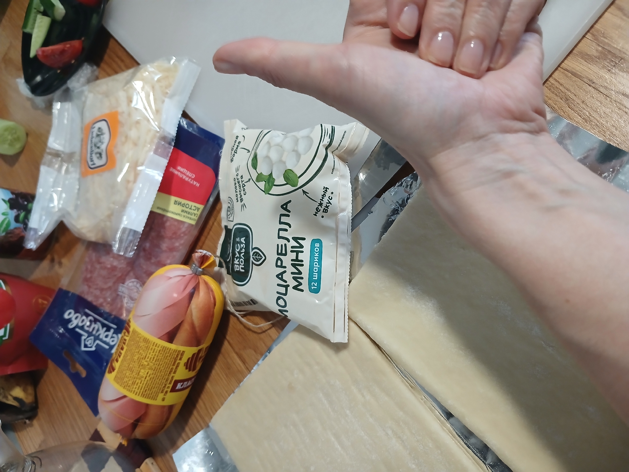 A close-up, high-angle shot shows a hand giving a thumbs-up over a collection of food items on a wooden surface. The items include a package of mozzarella balls, a vacuum-sealed package of sliced sausage, and what appears to be dough sheets. The background includes glimpses of other food items and packaging, suggesting a kitchen or food preparation area. The hand, belonging to an unidentified individual, is positioned in the foreground, obscuring a portion of the food. The lighting is natural and appears to be coming from overhead. The image provides no specific location in Yekaterinburg, Russia, or any identifying details beyond the visible objects.