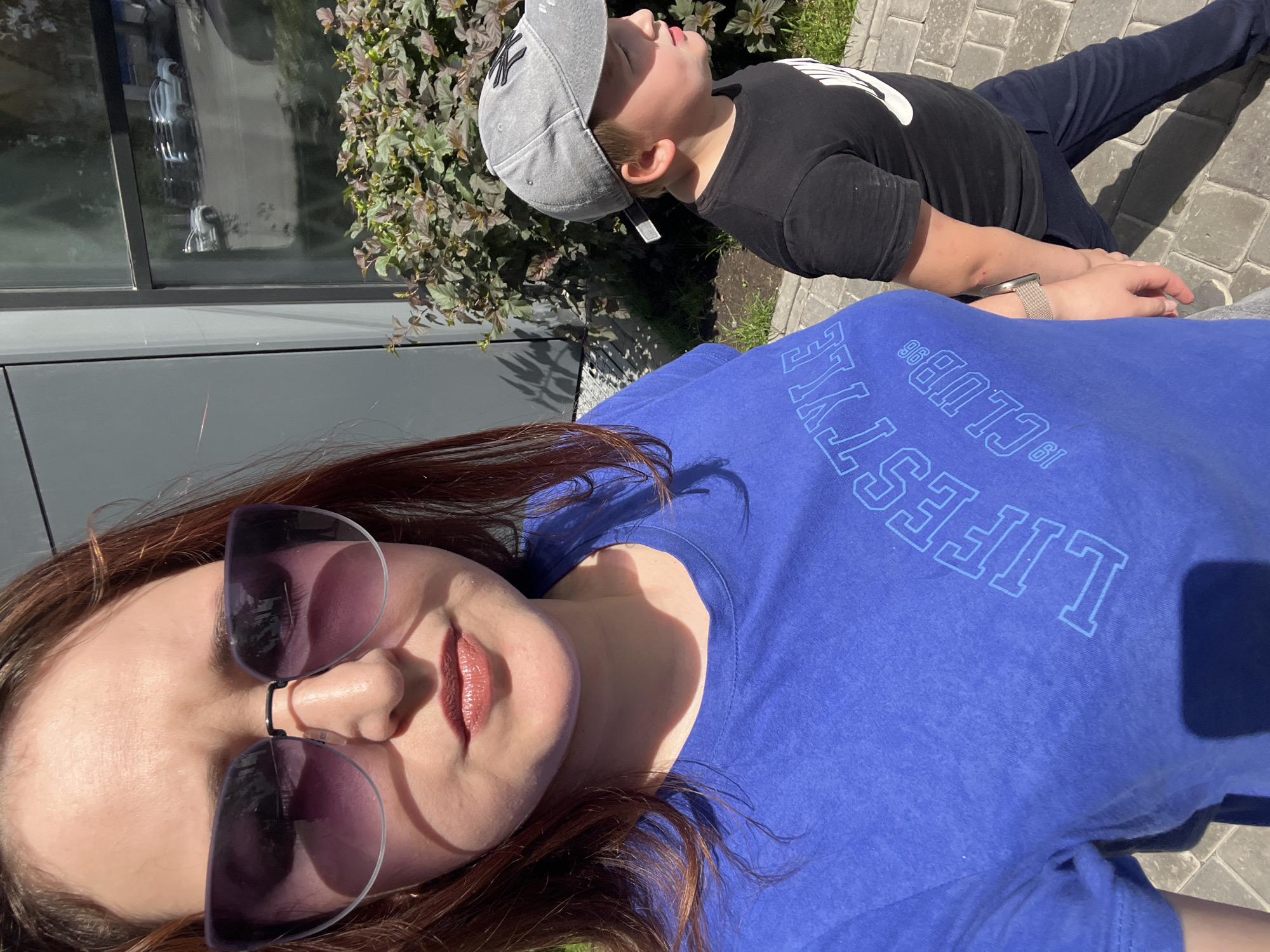 In a selfie taken outdoors, a woman with shoulder-length brown hair and sunglasses is visible from the chest up. She is wearing a blue t-shirt with the words "LIFESTYLE CLUB 96" printed on it in a lighter blue, outlined font. To her right, a young boy wearing a grey baseball cap and a black t-shirt with a white logo is walking away from the camera, holding the woman's hand. They are walking on a paved sidewalk. Behind them, there is a building with a large window reflecting cars and greenery, and a bush with dark green and reddish-purple leaves. The overall scene suggests a sunny day in an urban or suburban environment, possibly in Chelyabinsk, Russia, though this specific location cannot be confirmed from the visual information alone.