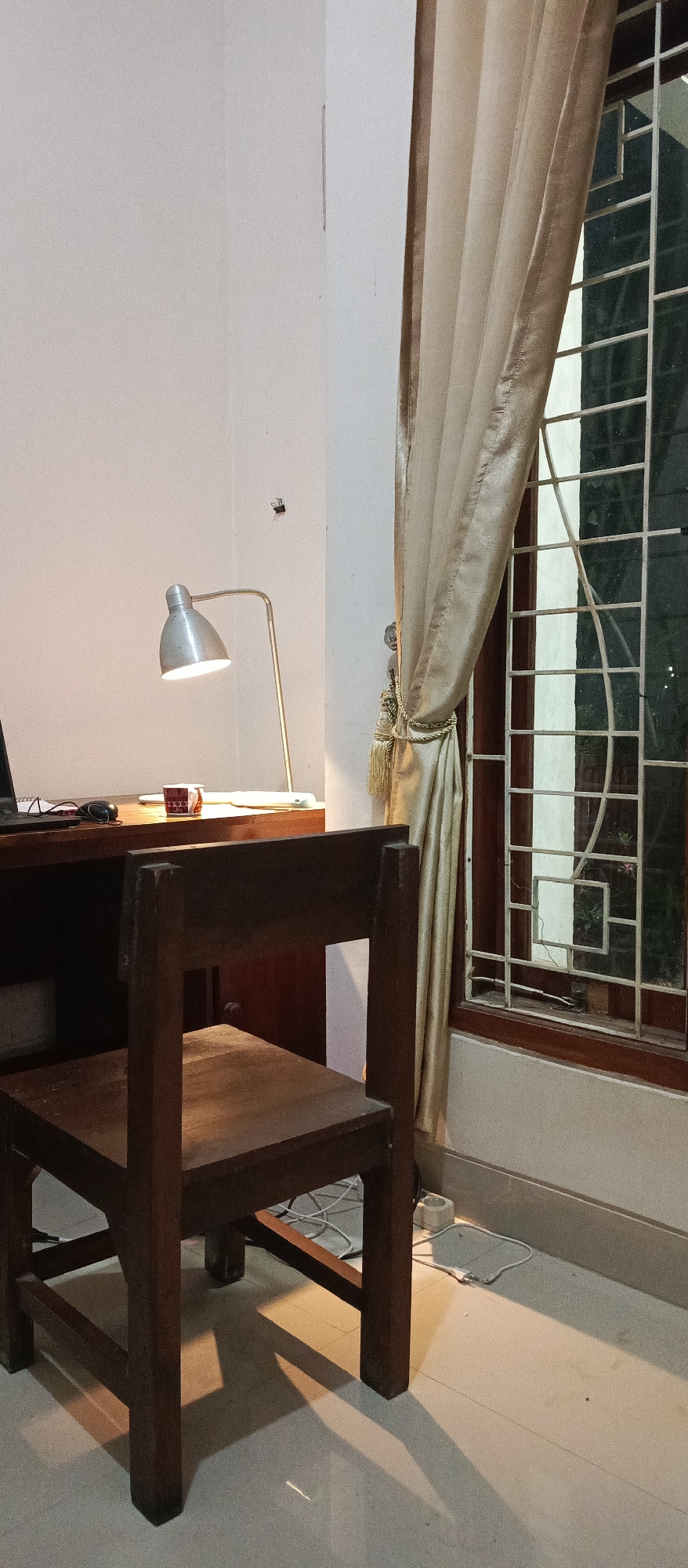 The image shows a desk with a lamp and a laptop, a wooden chair, a window with a patterned grate and curtains, and a white wall. A small object, possibly a spider, is visible on the wall. The scene appears to be an interior space, likely a room, given the presence of furniture and a window. The lighting suggests it may be evening or night. There are no people present. The overall impression is one of a simple, functional workspace. The location is unknown, and there are no discernible clues to suggest a specific city.