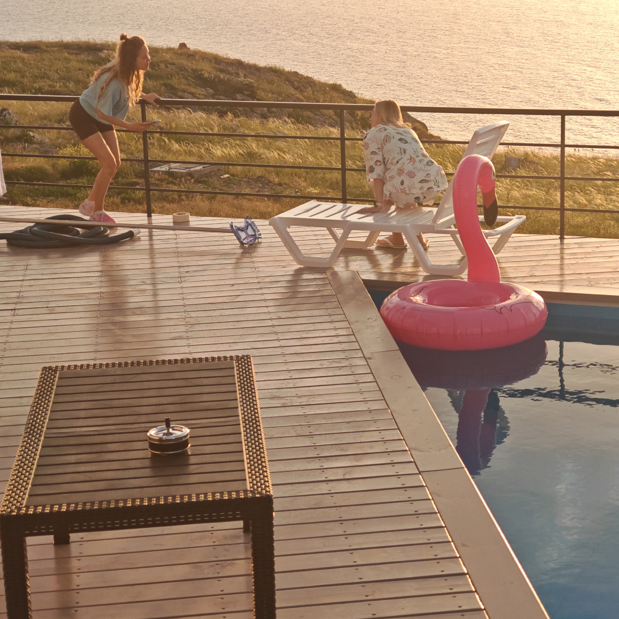 Here is a description of the image:
The image depicts an outdoor poolside scene in Кудьма, Russia, during golden hour. A wooden deck surrounds a pool with a pink flamingo inflatable floating in the water. Two women are visible on the deck: one stands near a railing looking out towards the sea, while the other is perched on a white lounge chair. A small dark table with an ashtray is in the foreground. Other items on the deck include a pool vacuum hose, a roll of tape, and a pool skimmer. The sea and a grassy, rocky hillside are visible in the background, with the sun reflecting on the water.