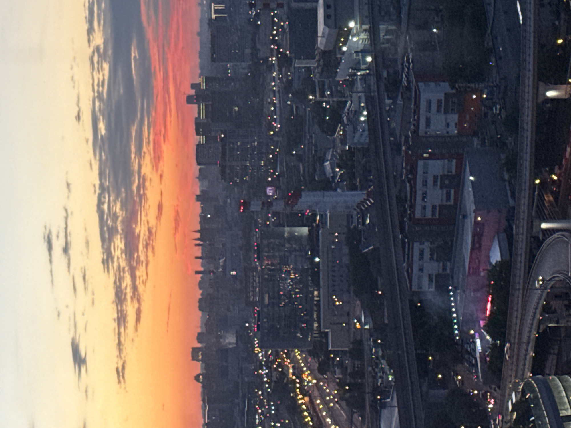 This is an aerial, nighttime photograph of the city of Khimki, Russia, likely taken during sunset or twilight. The sky is a vibrant mix of orange, red, and yellow hues, with scattered dark clouds. Below the sky, a sprawling urban landscape is visible, characterized by numerous buildings of varying heights and architectural styles.
In the foreground, several elevated roadways and highway overpasses curve through the scene, illuminated by streetlights and the headlights of passing vehicles. These roads are complex and multi-layered, indicating a busy transportation hub.
The mid-ground features a dense collection of buildings, including what appear to be commercial structures, residential complexes, and industrial facilities. Many of these buildings have illuminated windows, and some display prominent red and white lighting, possibly from signage or architectural features. One notable structure in the mid-ground is a tall, cylindrical building with red and white bands. Another large building, appearing to be a modern office building or shopping center, is visible with numerous lights reflecting from its facade.
The background showcases a more distant skyline of taller buildings, some with illuminated tops. The overall impression is of a densely populated and economically active urban area at dusk, with the city lights beginning to dominate the fading natural light. The image captures the transition from day to night in a bustling Russian city.