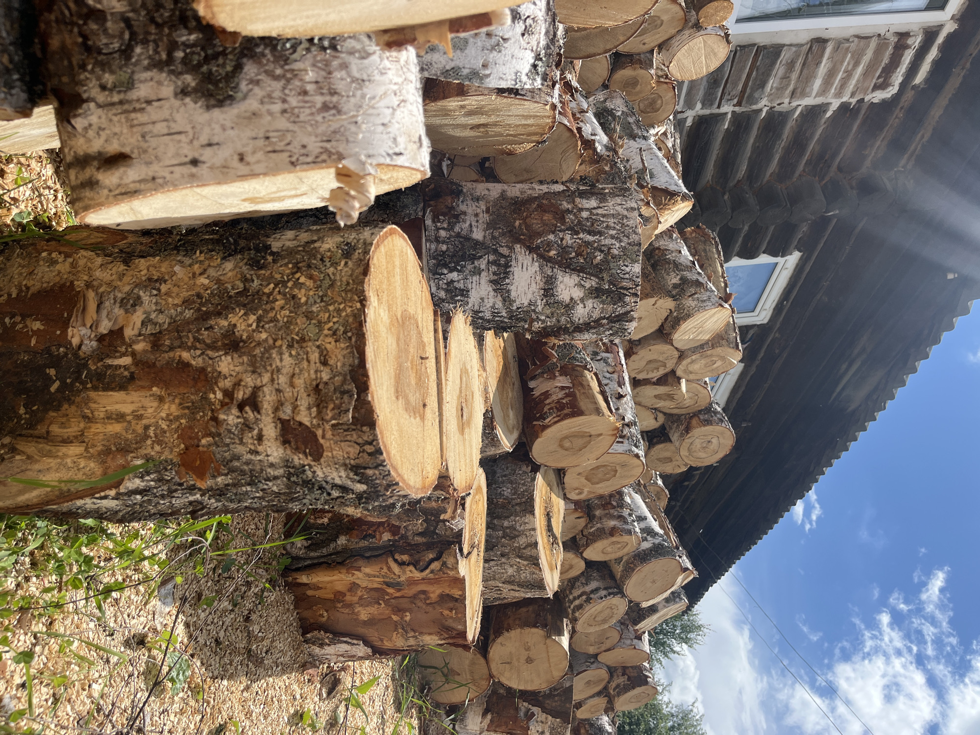 The image depicts a large pile of freshly cut logs stacked against the side of a rustic, wooden log cabin. The cabin appears to be made of dark, horizontal logs with some white material filling the gaps. It has a dark, corrugated roof and windows with white frames. The logs are of various sizes, some with visible bark, while others are smooth and show the concentric rings of the wood. The ground around the logs is covered with wood chips and some green grass. In the background, trees and a blue sky with scattered white clouds are visible. The overall scene suggests a rural setting, possibly in or near the city of Уральское, Russia, given the prompt. There are no people or animals present in the image.