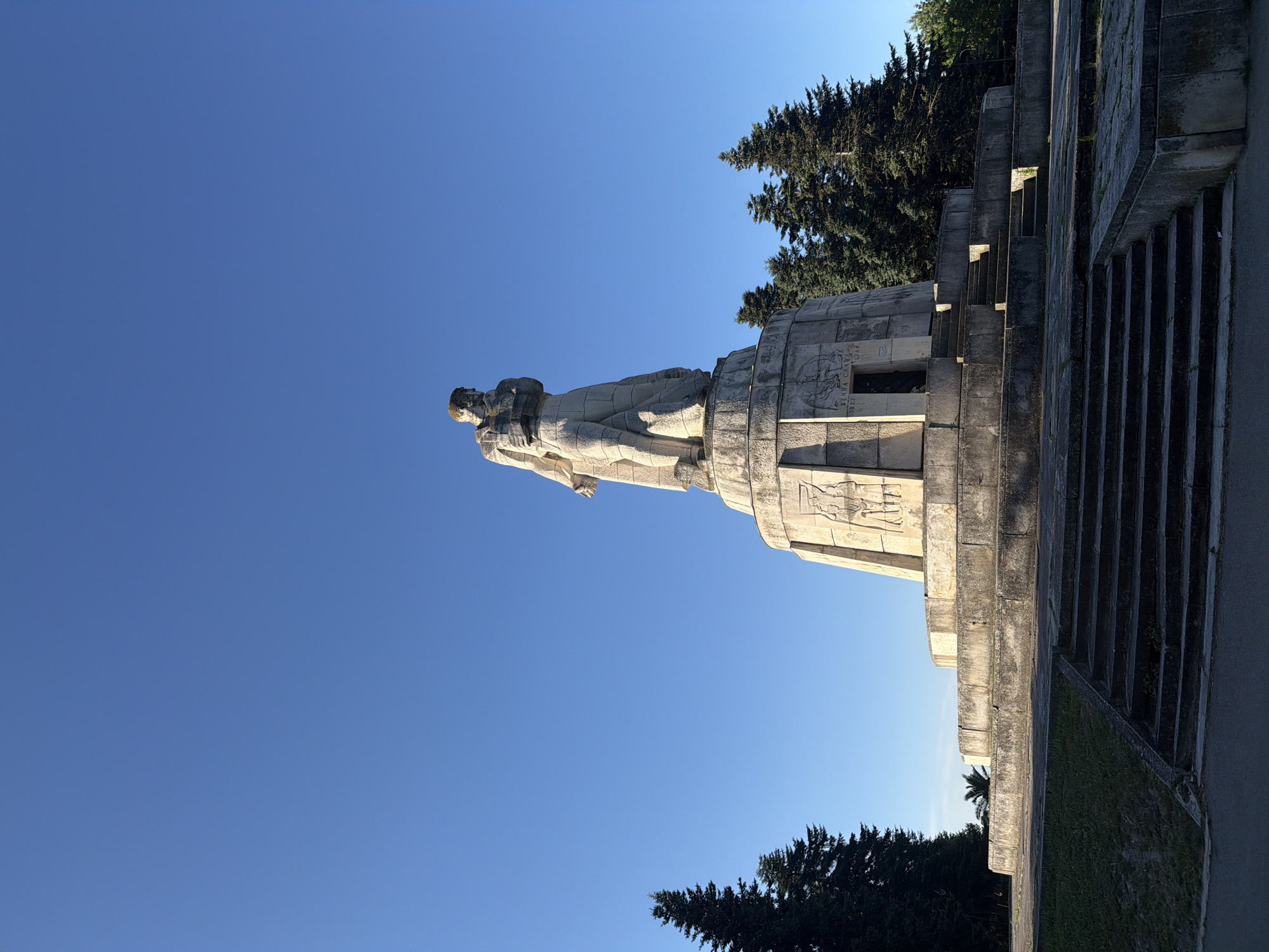 The image depicts the Monument of the Bulgarian-Soviet Friendship in Varna, Bulgaria. It is a monumental statue made of stone, representing two figures, a woman and a man, with the man embracing the woman. The statue stands on a large, tiered concrete pedestal. The pedestal is decorated with bas-reliefs depicting scenes of historical significance and text in Cyrillic script.
The monument is situated in an open area with a clear blue sky in the background. Surrounding the monument are several tall, dark green pine trees. In the foreground, there are concrete steps leading up to the monument and a grassy area. The overall scene is one of a grand, somewhat imposing, public monument under a bright sky. The lighting suggests it is daytime, likely morning or afternoon due to the clear, crisp shadows.