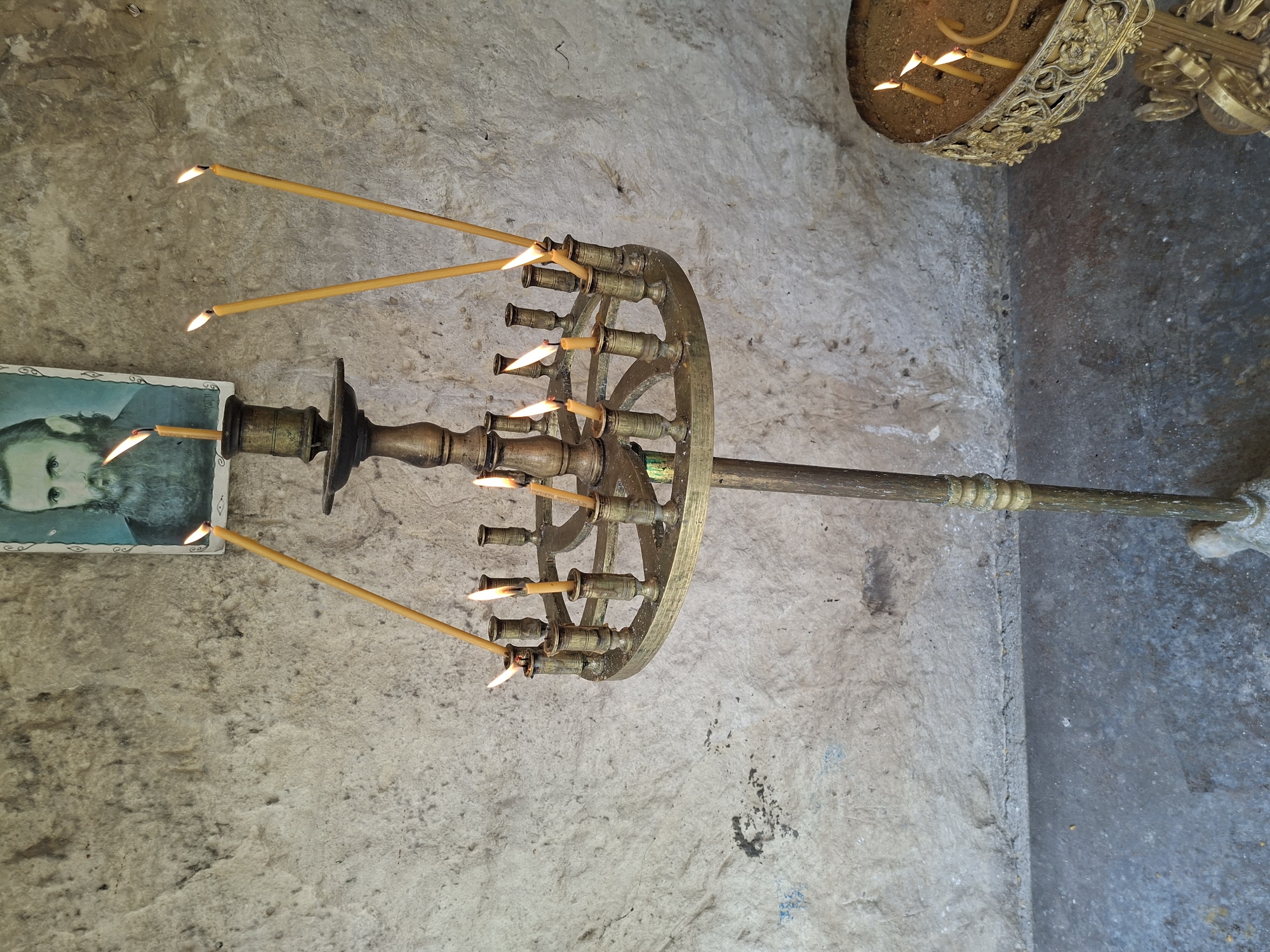 The image captures an interior view within a religious setting, identified as Basarbovo, Bulgaria. Dominating the foreground is a tall, ornate brass candelabra featuring a central stem and a circular lower tier. Multiple thin, yellow wax candles are actively burning on this candelabra; four longer candles extend upwards and outwards, while several shorter ones are positioned within the circular tier's holders. To the upper left, a framed portrait is affixed to a roughly textured, light-grey stone or rock wall. The portrait depicts a bearded man with a somber expression, possibly a cleric or saint, rendered in an iconic style with a light-colored, decorative frame. The wall exhibits irregularities, darker patches, and some faint blue markings. In the lower right corner, a second, smaller, and more ornate gilded or brass candle stand is visible. Its top features a bowl-like depression filled with sand, holding at least three lit, shorter yellow candles. The floor beneath the candelabra is a dark, worn concrete surface, meeting the textured wall at a distinct line. The overall scene suggests a devotional area, possibly within a monastery or church.