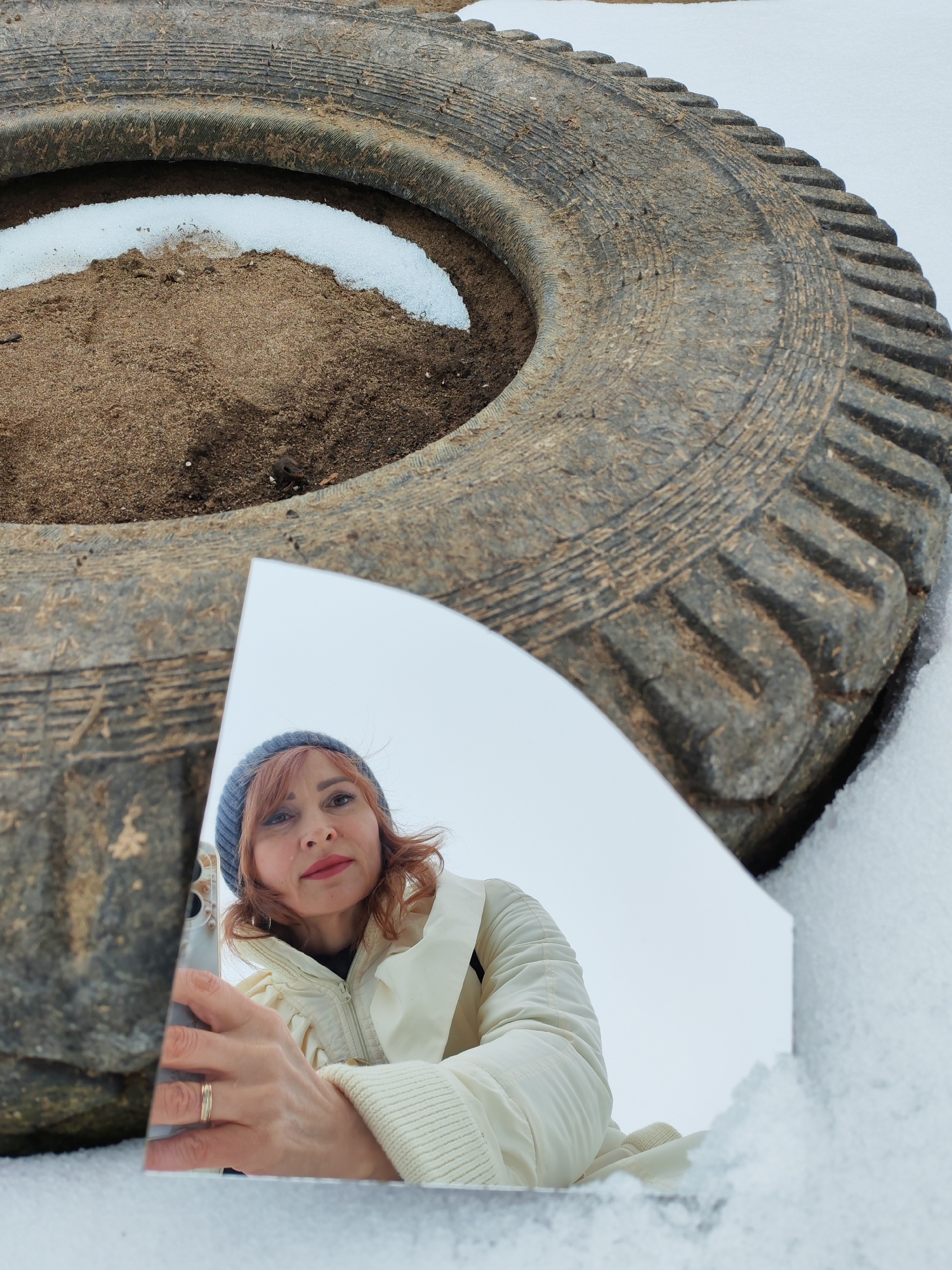 A woman is reflected in a shard of mirror held up in front of a large, old tire in a snowy landscape. The tire is filled with dirt and some snow. The woman has red hair and is wearing a blue beanie and a white jacket. The reflection shows her holding a phone and looking at the camera. The background is snowy. The tire appears to be partially buried in snow. The location is Chaikovsky, Russia.