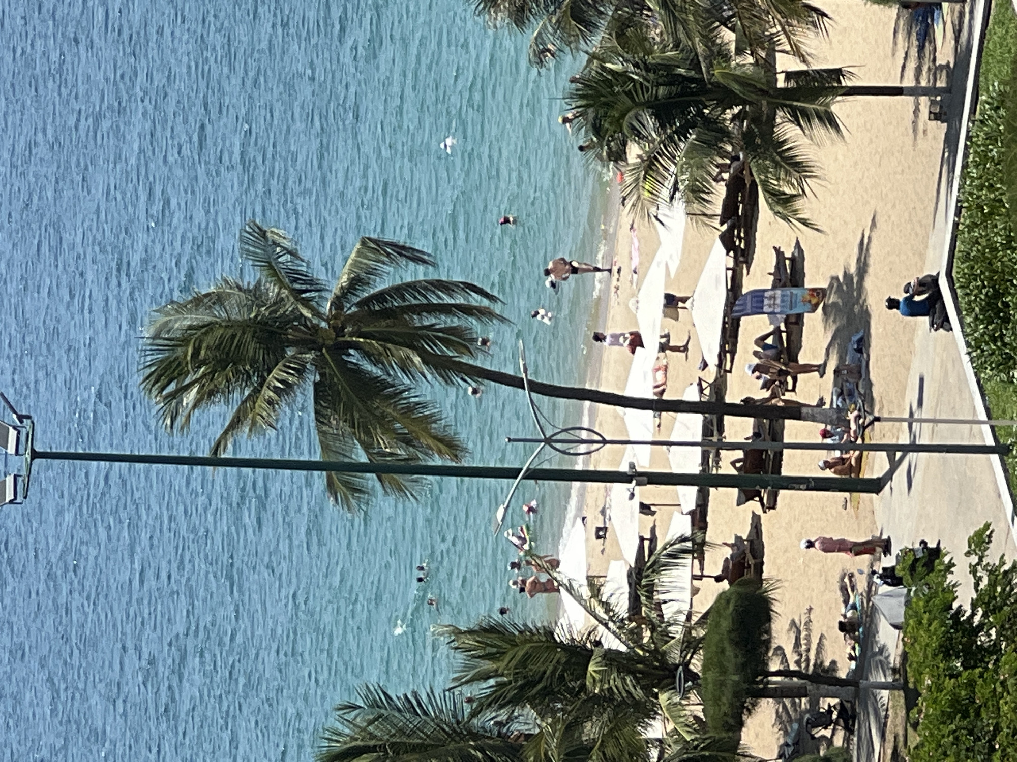 The image depicts a bustling beach scene in Nha Trang, Vietnam, characterized by the azure expanse of the ocean meeting a sun-drenched sandy shore. Palm trees, a quintessential element of tropical landscapes, are scattered throughout the frame, their fronds swaying gently in the breeze. Numerous beach umbrellas provide shade to beachgoers, some of whom are lounging on chairs or towels, while others are actively enjoying the water. Several individuals are visible swimming or wading in the shallow sea, their figures silhouetted against the shimmering surface. A prominent lamppost stands tall in the foreground, its metallic structure contrasting with the natural elements. The beach is populated by a diverse mix of people, engaged in various recreational activities, creating a vibrant and lively atmosphere. The scene is bathed in bright sunlight, suggesting a clear and pleasant day.