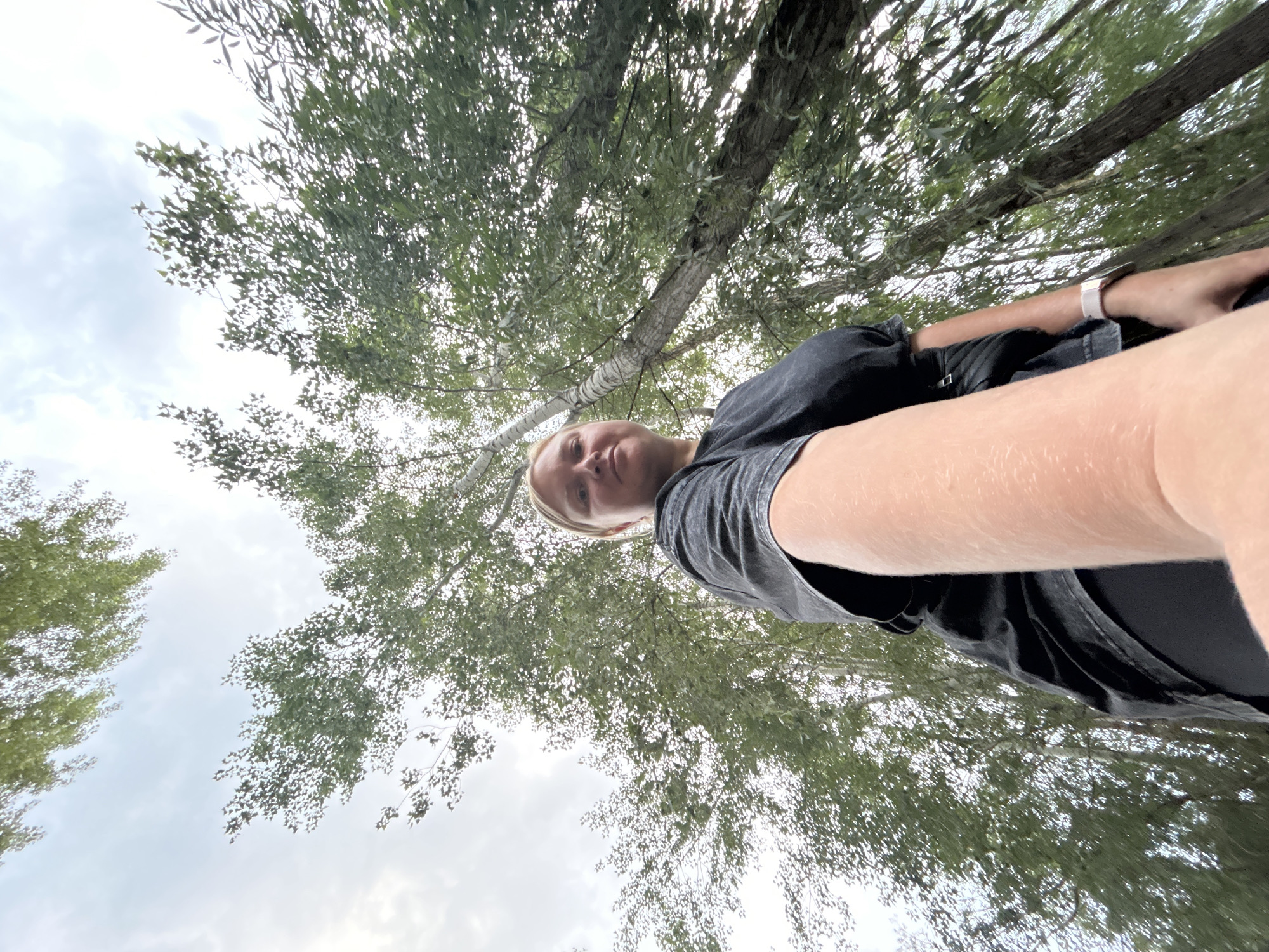 A low-angle, medium shot shows a woman taking a selfie outdoors amidst trees under an overcast sky. The woman, with blonde hair and fair skin, is wearing a dark t-shirt and dark pants. Her right arm is extended towards the camera, and her face is visible in the right portion of the frame, looking towards the viewer with a neutral expression. The background is filled with lush green foliage and tall tree trunks, suggesting a park or wooded area. The sky is cloudy, indicating a temperate day. The scene is devoid of any specific landmarks that would pinpoint its location within Chaikovsky, Russia.