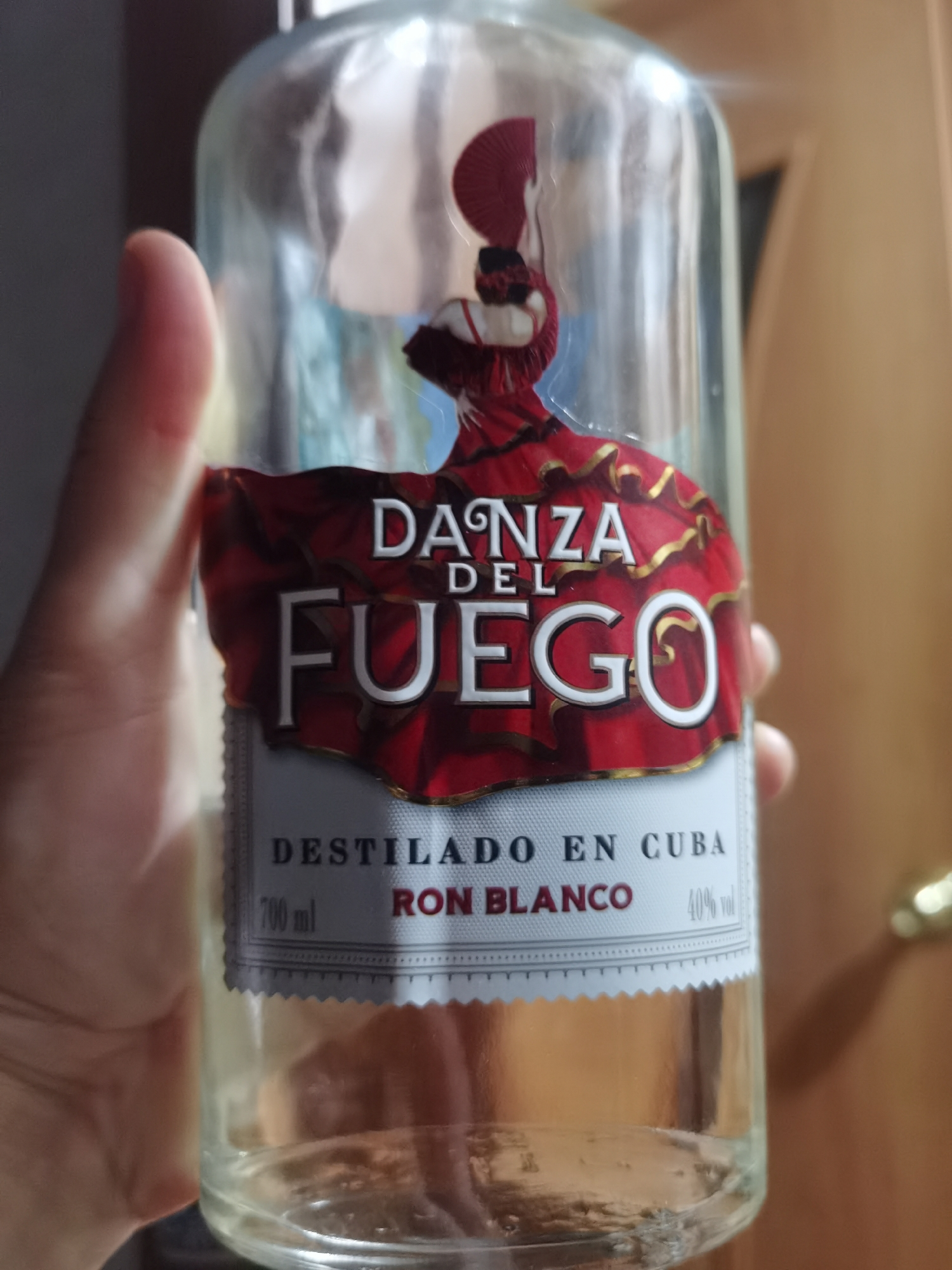 A clear glass bottle of rum, labeled "Danza del Fuego" and "Ron Blanco", is held in a person's hand in front of a blurred background. The bottle is partially filled with a light-colored liquid. The label features a stylized illustration of a woman in a red dress with a fan. The background appears to be an indoor setting with a wooden door visible on the right. There is no indication of the city of Chaikovsky, Russia, or any other specific city in the image.