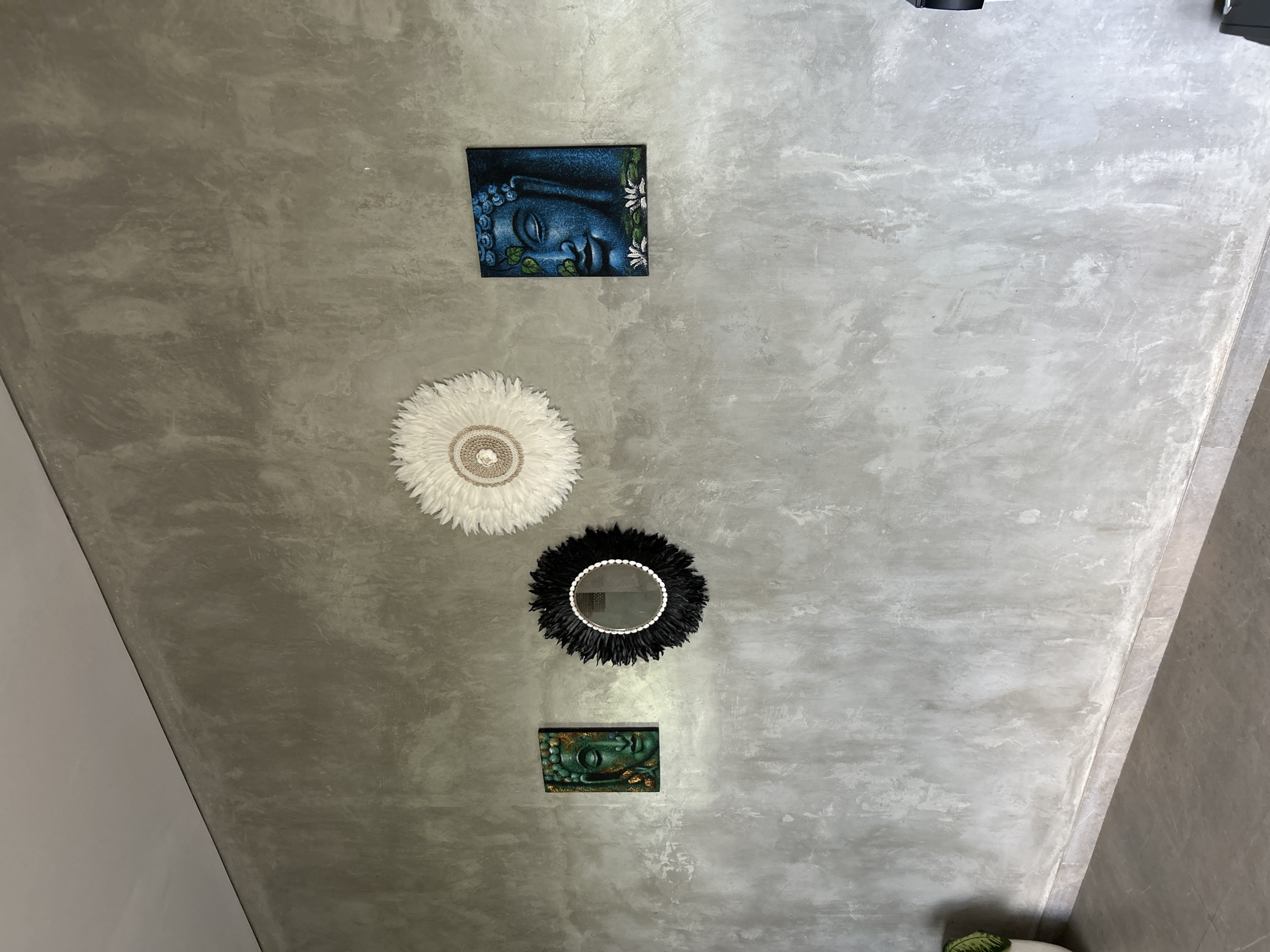 The image shows a textured gray wall in what appears to be a room. Mounted on the wall are three decorative items. On the left is a rectangular painting of a green Buddha face with gold accents. To its right is a round mirror with a black, spiky frame, possibly made of feathers or similar material, and a border of small white beads. Further to the right is a circular wall decoration made of white feathers, with a woven center. To the far right, a second rectangular painting depicts a blue Buddha face with green leaves and white water lilies. A small portion of a green plant in a pot is visible in the bottom left corner. The floor appears to be tiled. The scene is static and devoid of any people or actions. The setting is identified as Tembuku, Indonesia.