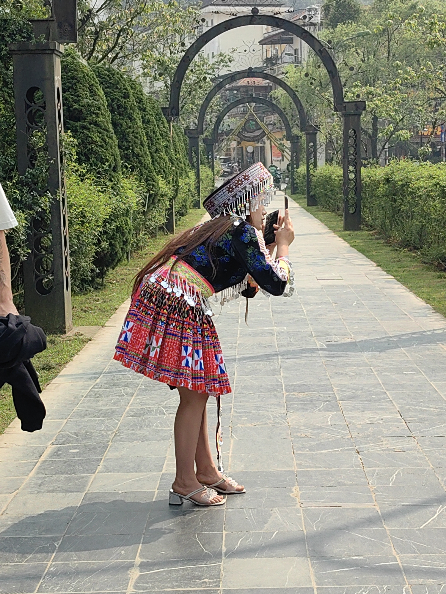 The scene is an outdoor paved pathway in Sa Pa, Vietnam, under bright daylight. A person in traditional Hmong attire is positioned centrally, bending forward at the waist, holding a black smartphone horizontally with both hands as if photographing. This individual wears a silver-decorated headpiece, a dark embroidered long-sleeved top, a vibrant pleated skirt with red, white, blue, and black geometric patterns, and silver-colored coin embellishments. Light-colored block-heel sandals are on her feet. To the left, a partial view of another person shows a left arm in a white t-shirt holding a dark garment. The pathway, constructed of grey rectangular slabs, is flanked on the left by a tall, manicured green hedge and on the right by a series of dark, ornate metal arches with decorative pillars, alongside lower green shrubs. Light-colored urban buildings are visible in the distant background, partially obscured by trees.