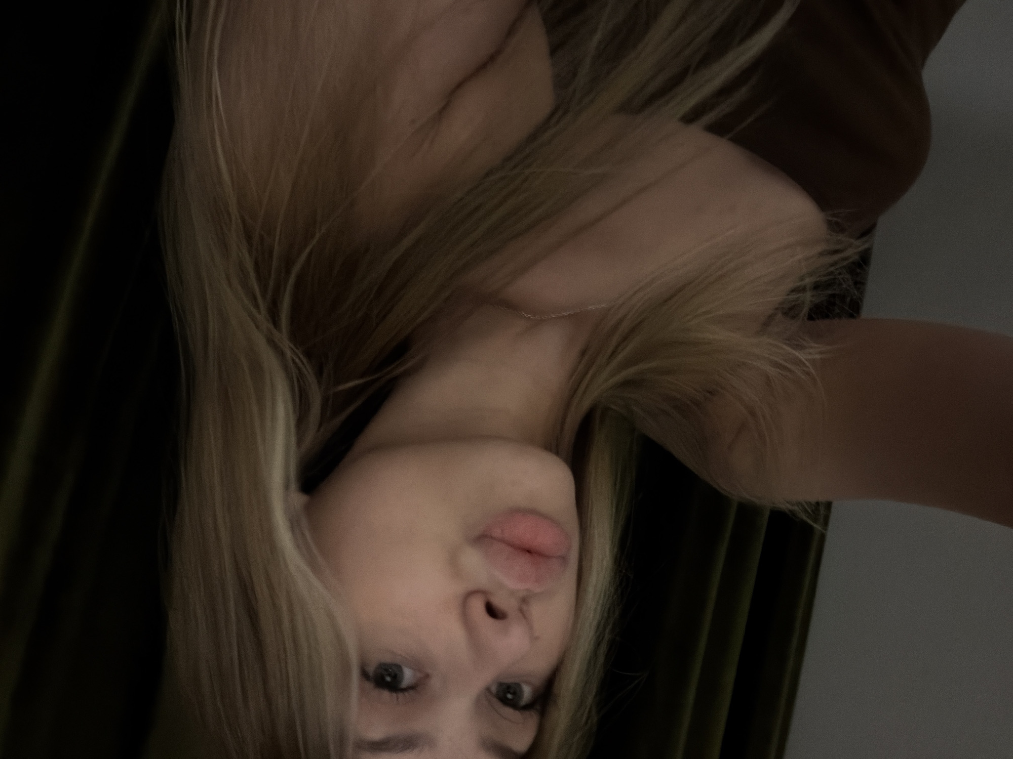 A young woman with blonde hair and light skin is pictured from the chest up, looking towards the camera. She is wearing a brown strapless top and a thin necklace. Her lips are slightly parted in a pout. Her blonde hair is long and reaches her shoulders, with some strands falling across her face and chest. The background is dark, with what appears to be a dark green curtain on the right side of the image. The lighting is dim, creating a shadow on the left side of her face. The scene does not provide any specific details to indicate a location in the city of Đà Nẵng, Vietnam.