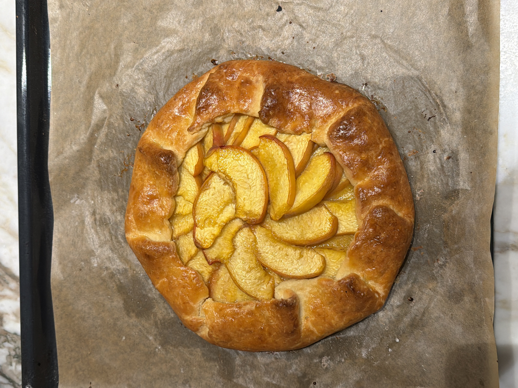 A close-up, overhead shot shows a golden-brown peach galette on a baking sheet lined with parchment paper. The galette has a flaky, crimped crust folded over sliced, caramelized peaches. The baking sheet sits on a marble countertop. The image is lit from above, casting subtle shadows.
