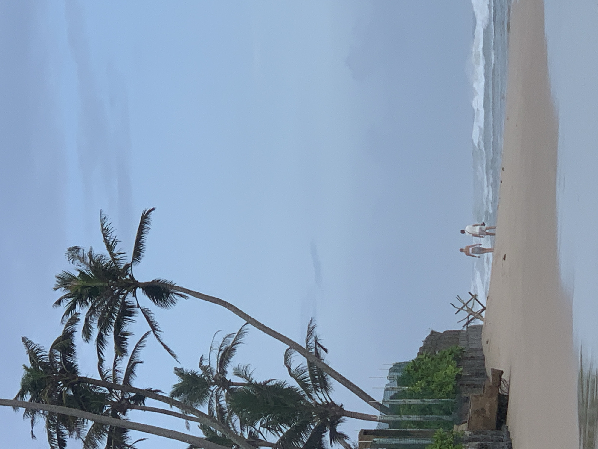 The image depicts a beach scene in Kathaluwa, Sri Lanka. The beach is sandy, and waves are breaking on the shore. Two people are walking along the beach. In the foreground, there are several palm trees leaning towards the right. Behind the palm trees is a building structure with greenery. The sky is overcast.